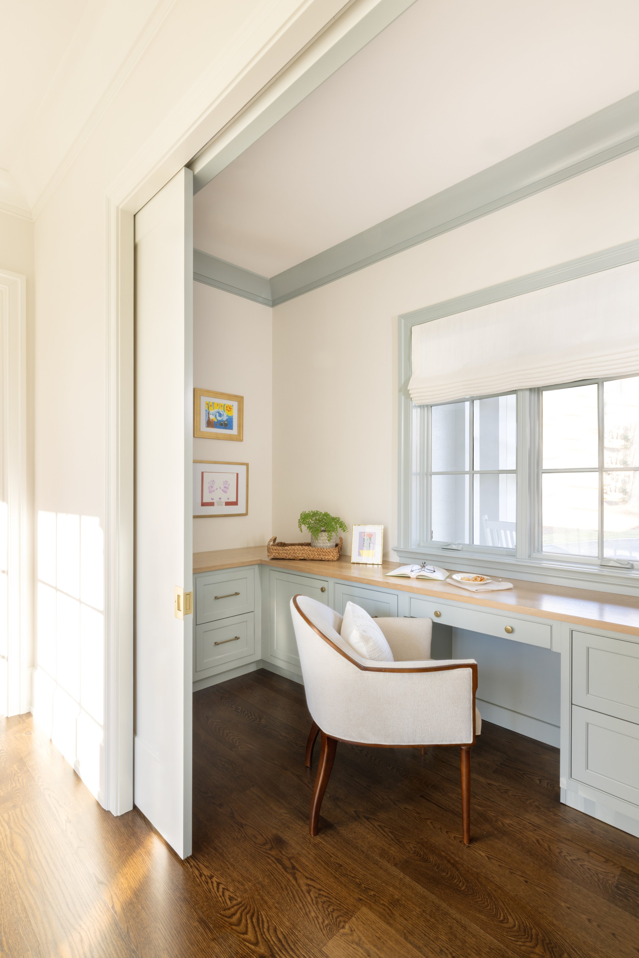 A cozy corner of a room with a built-in desk by a window, a cream-colored chair with wooden legs, a small potted plant, and framed artwork on the wall.