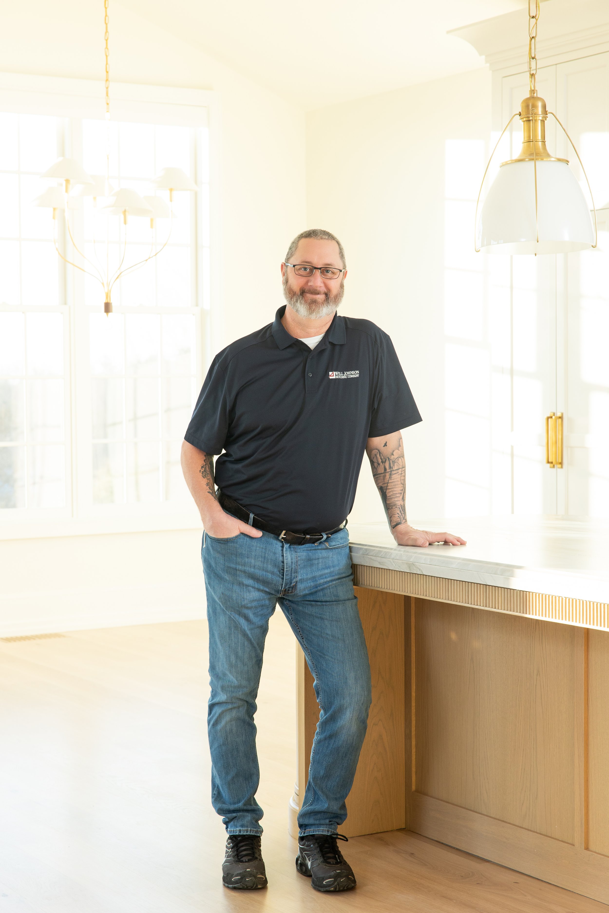Man wearing a cap and polo shirt reviewing architectural blueprints on a table, with a ruler and notebook nearby, in a room with built-in shelves and a television.