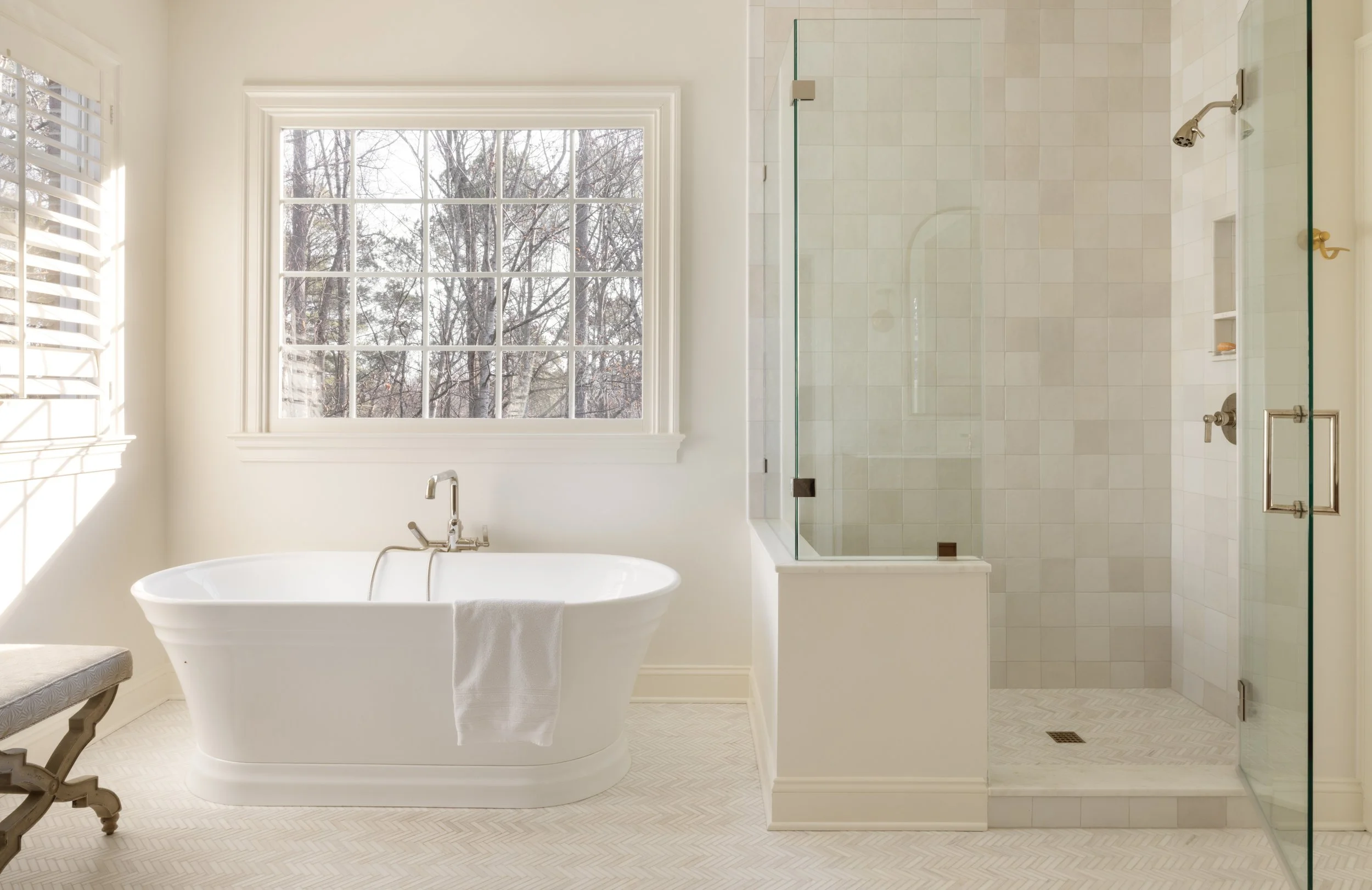 A bright bathroom with a large window, a white bathtub, and a glass-enclosed shower with tiled walls.