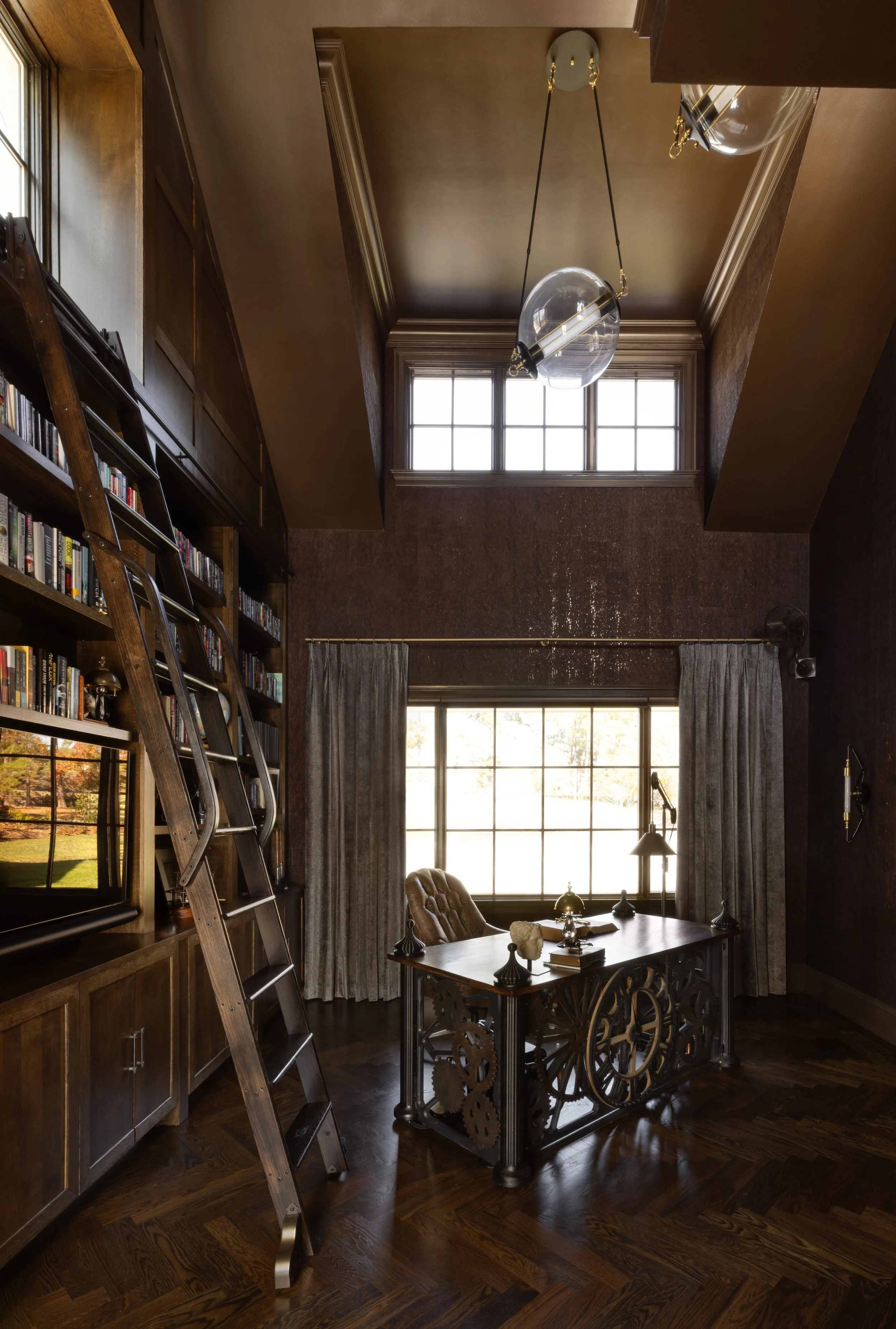 A home office with dark wood bookshelves, a ladder, a large window with gray curtains, a wooden desk with decorative gears on its sides, and a tufted leather chair, illuminated by natural light.