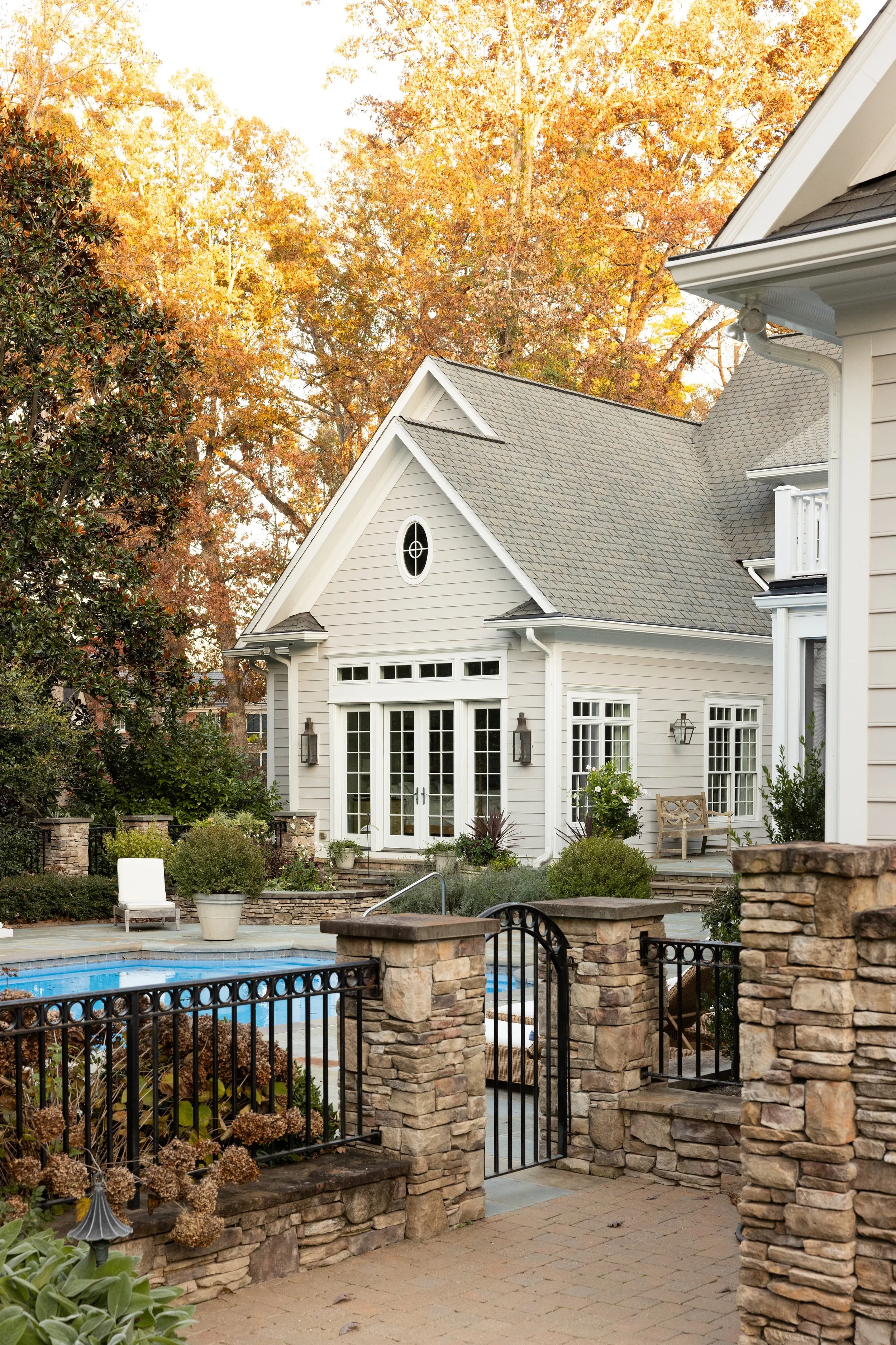 Residential house with a backyard featuring a swimming pool, surrounded by stone and metal fencing, with plants and trees with fall foliage.