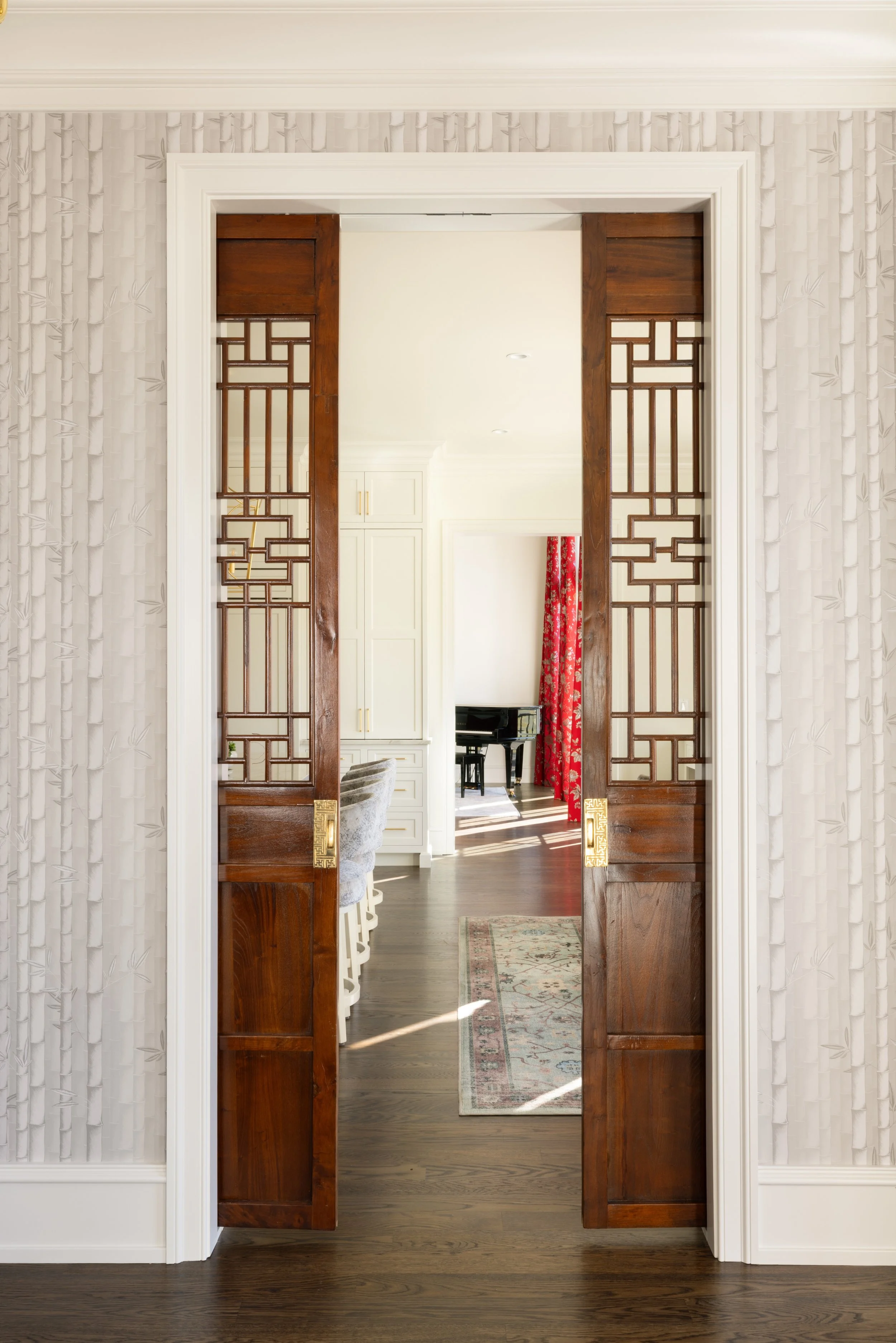 Interior view of a home with a pair of wooden sliding doors with decorative geometric patterns, opening into a living space with a black grand piano, white cabinetry, a patterned rug, a red floral curtain, and hardwood floors.