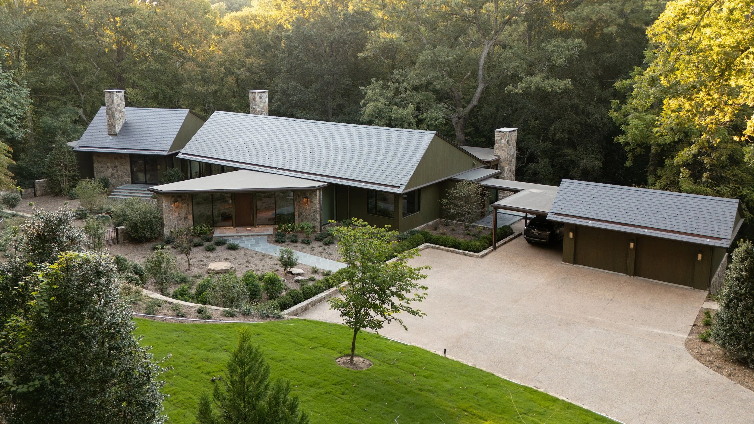 A modern house with multiple gable roofs, stone and green exterior walls, surrounded by trees and landscaped yard.