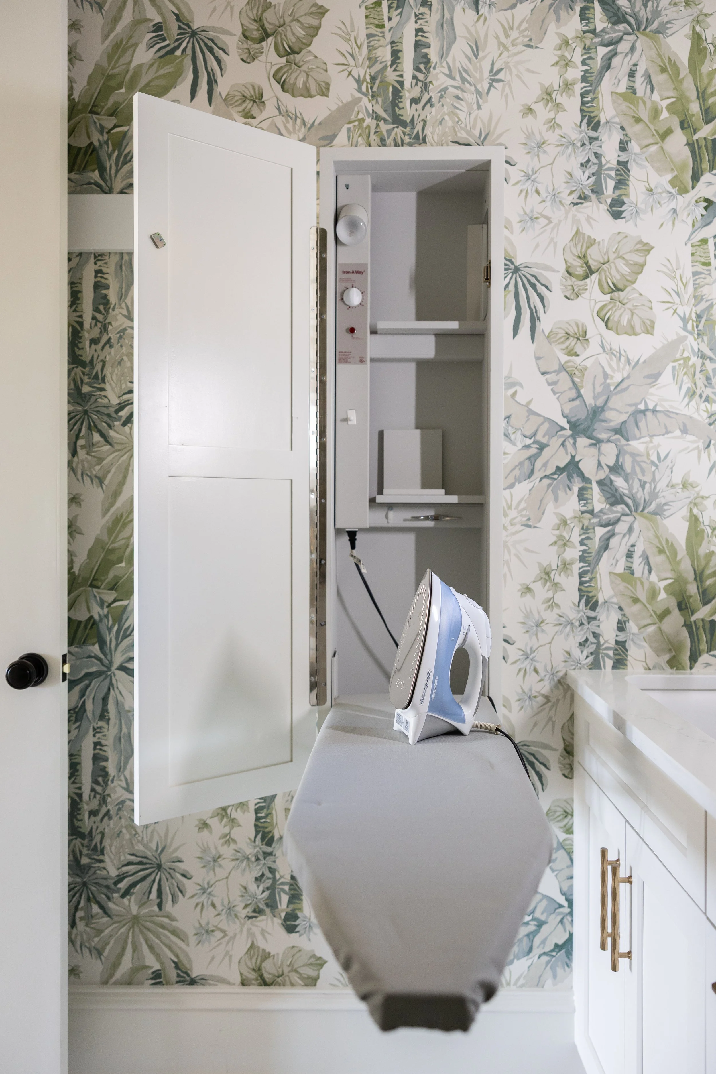 A laundry room with botanical wallpaper, a cabinet with an open door, an ironing board with an iron, and a utility shelf with an iron on it.
