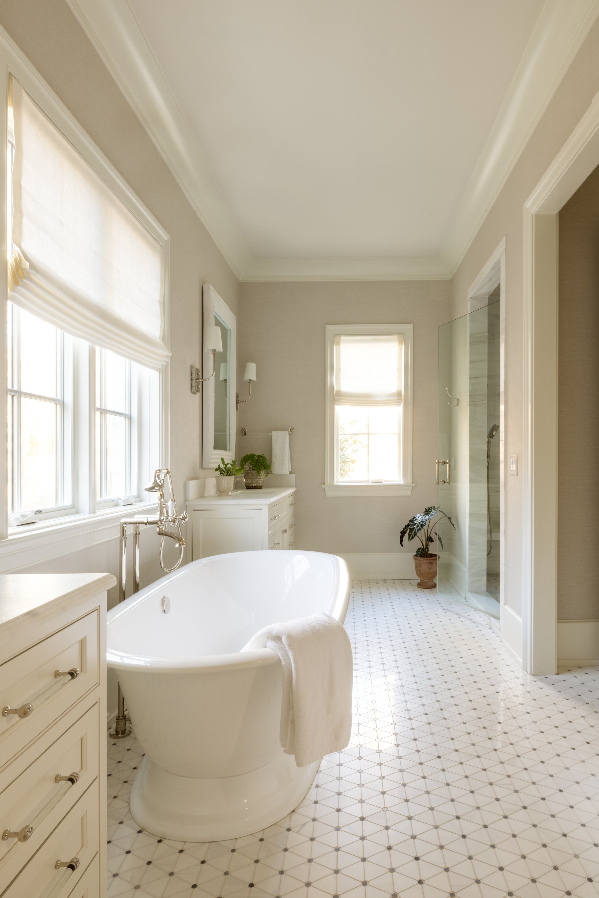 Bright bathroom with a white bathtub, large windows with roller shades, a white vanity, and a glass-enclosed shower. Decor includes potted plants, a mirror, and wall-mounted lights.