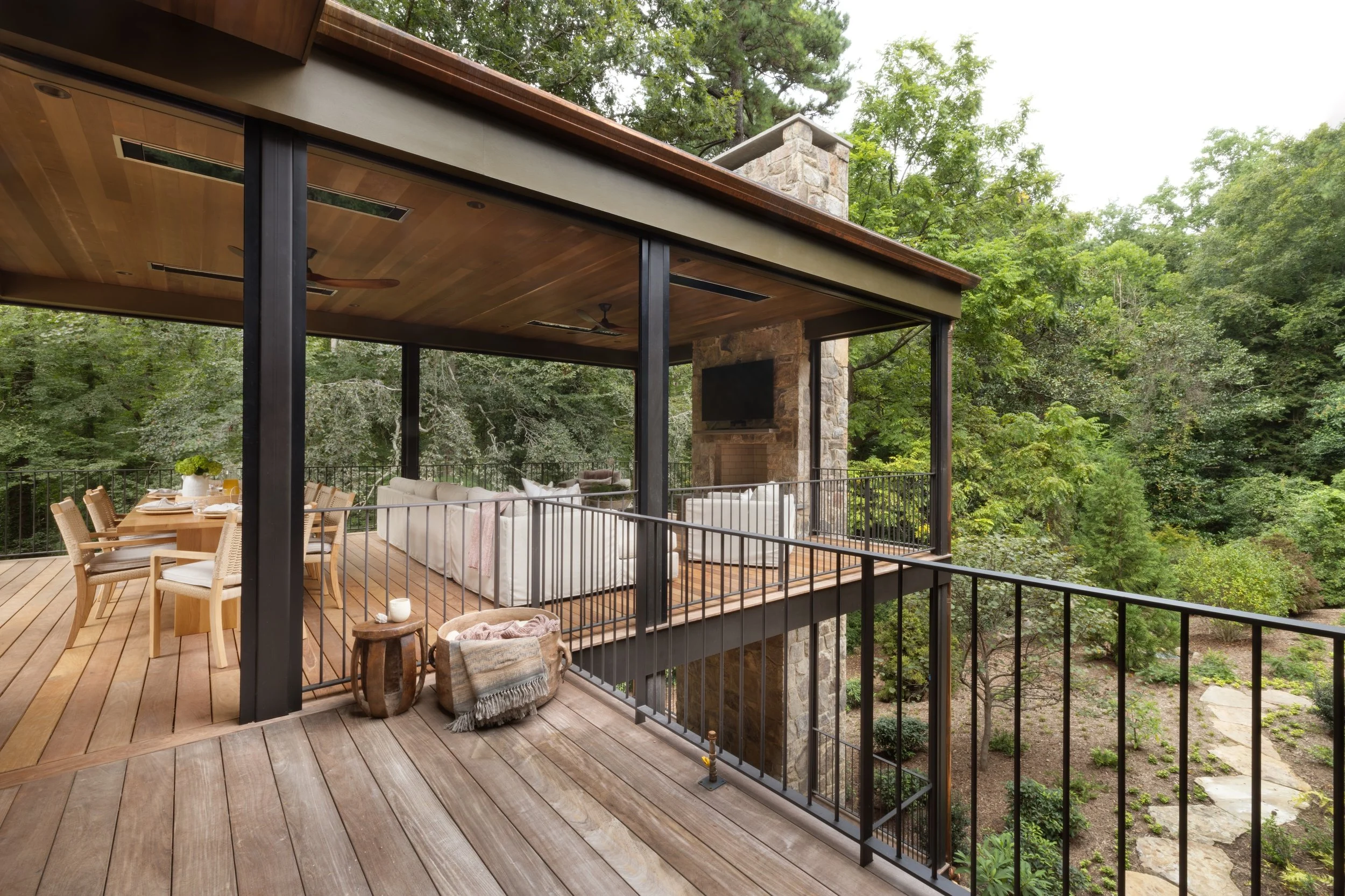 Outdoor deck area with wooden flooring, black metal railing, ceiling fan, and stone fireplace with mounted TV, surrounded by lush green trees.
