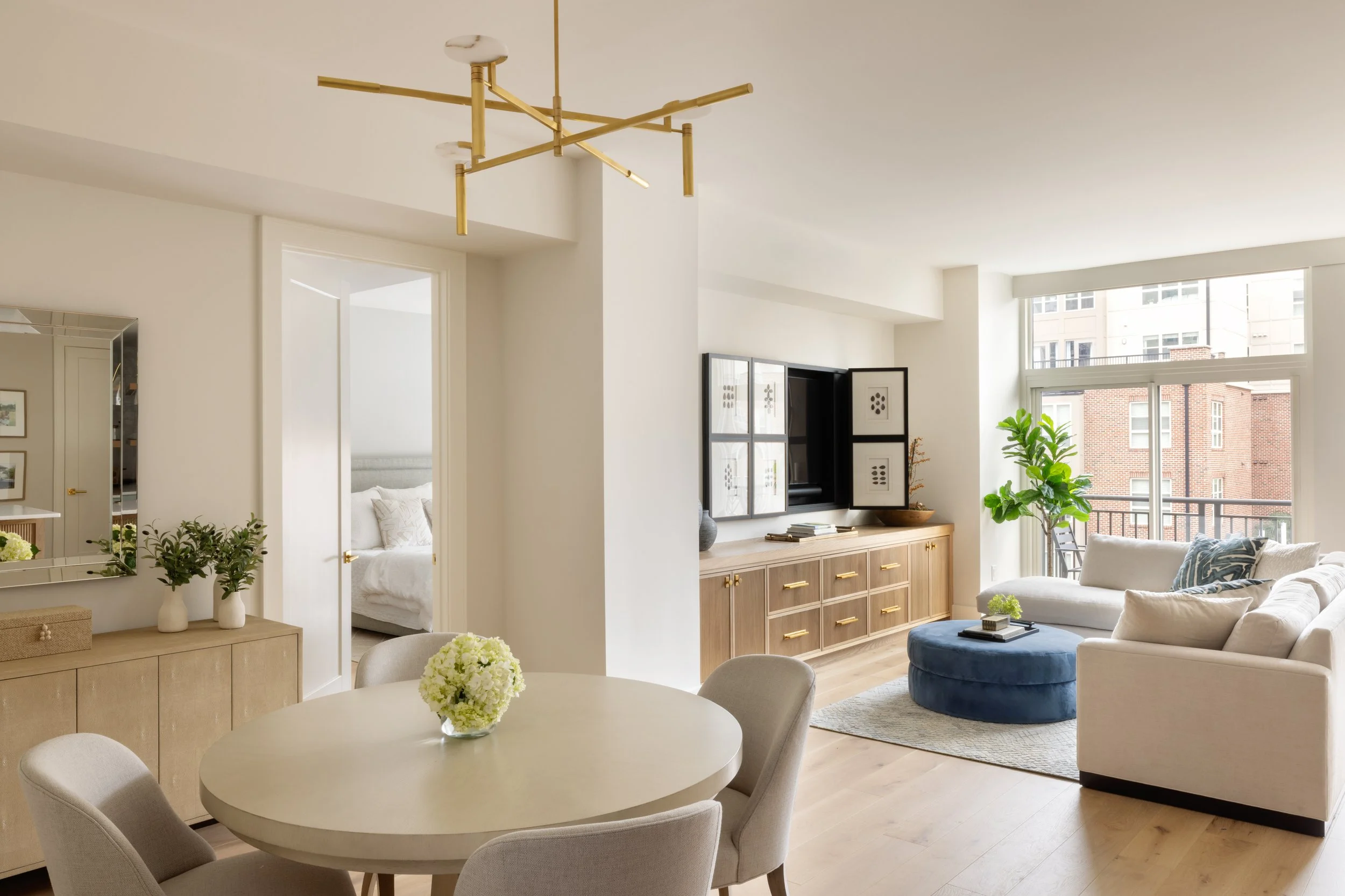 Bright living room with cream-colored sofas, a blue ottoman, large window, and wooden furniture, including a sideboard and a dining table with chairs, decorated with green plants and flowers.