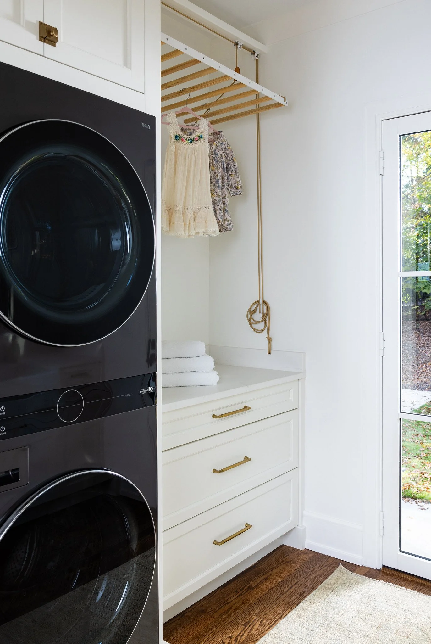 A laundry room with a black front-loading washing machine, white cabinet with three drawers and gold handles, a stack of white towels, hanging dresses, and a side door with a view of a yard outside.