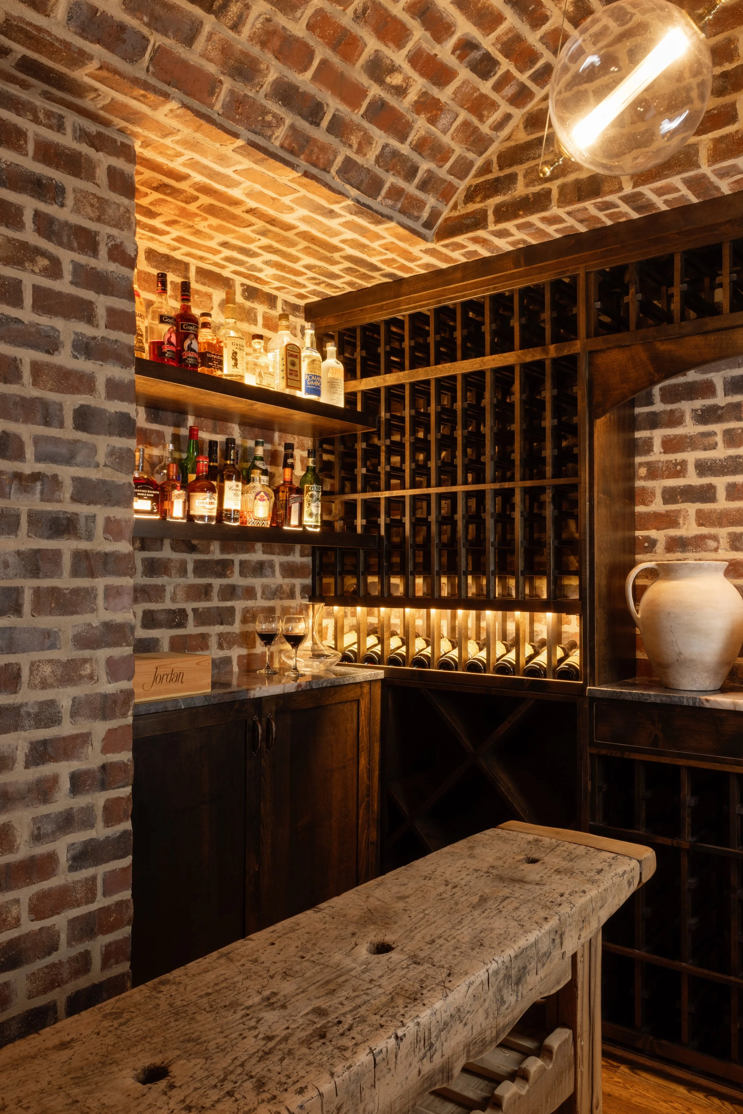 A cozy wine cellar with brick walls, wooden shelves filled with liquor bottles, a wine rack with bottles, glasses of red wine on a marble surface, and a rustic wooden table.