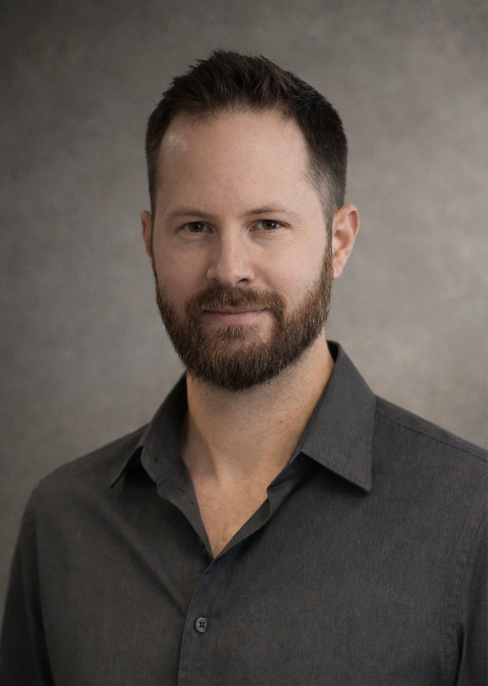 Portrait of a man with a beard and short dark hair, wearing a dark gray button-up shirt, against a neutral gray background.