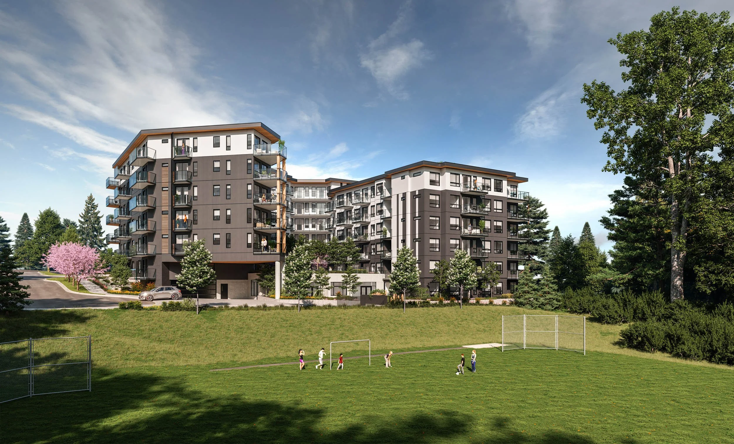Six-storey condominium seen across a grassy playing field where children are playing soccer, framed by tall evergreens