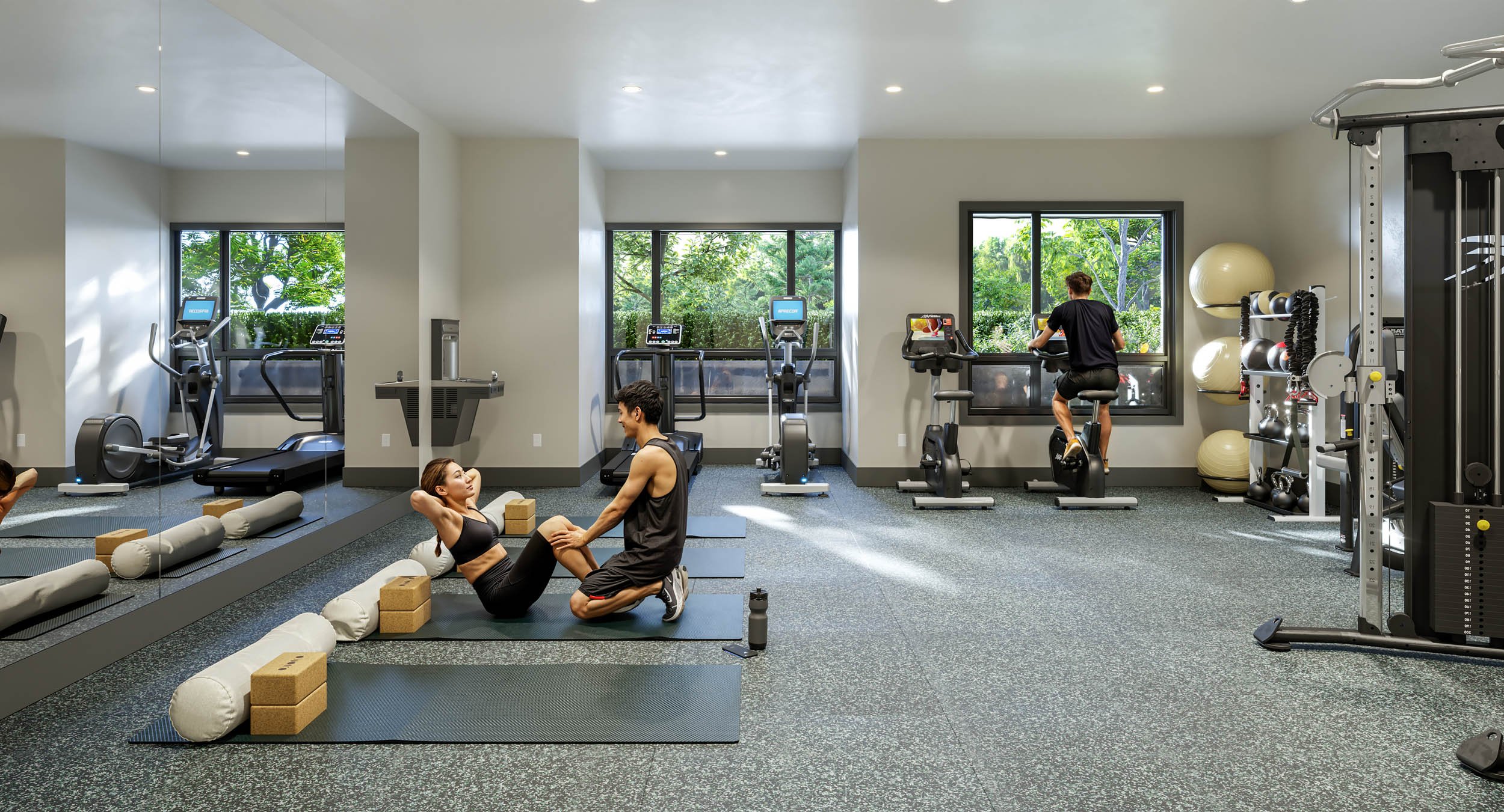 Two residents stretching on mats in a bright fitness room, with treadmills and exercise bikes along the windows, a mirrored wall and a rack of weights and stability balls