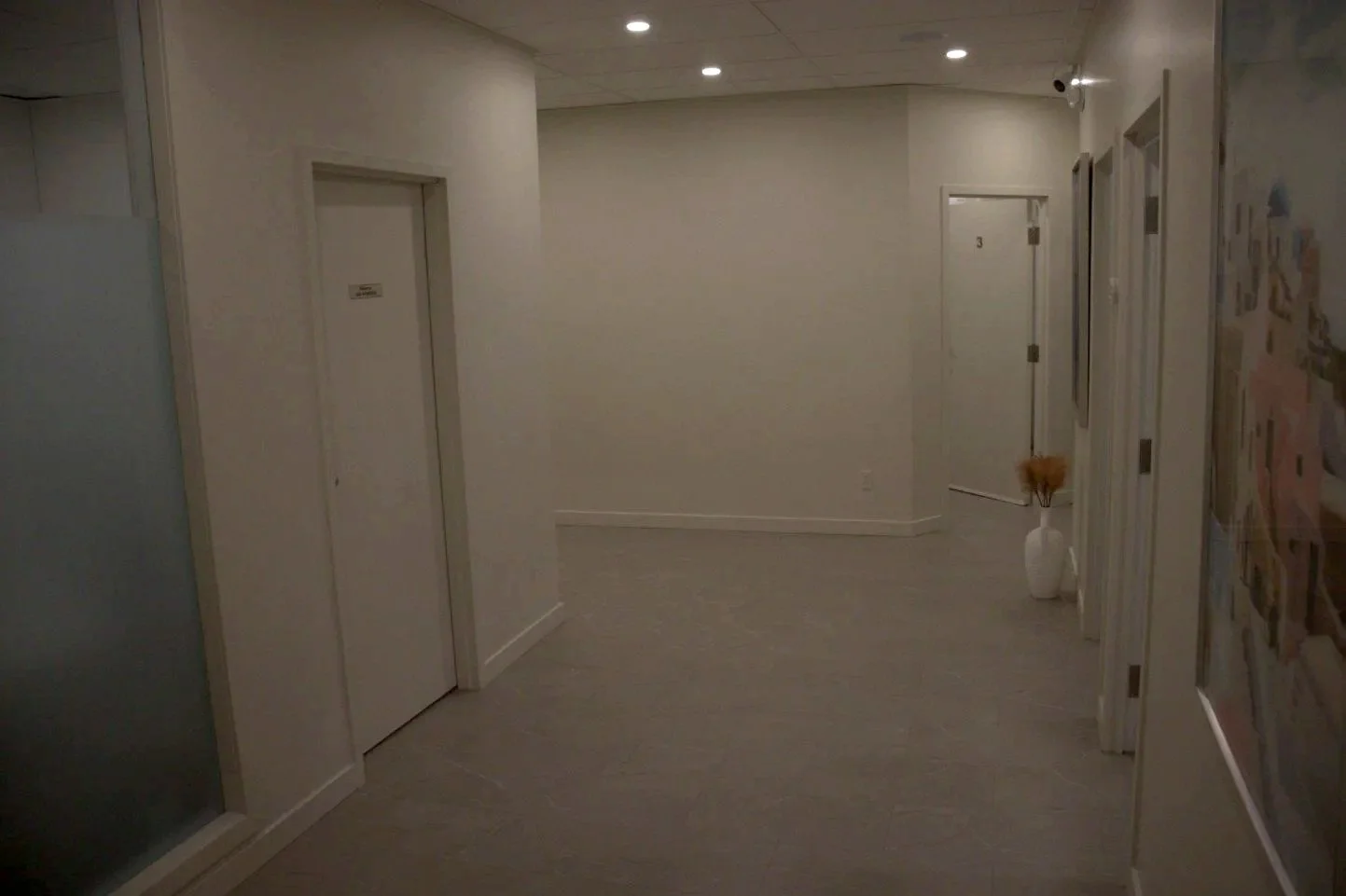 Un couloir dans un bâtiment avec deux portes d'ascenseur et une porte de chambre, un vase avec des fleurs en céramique blanche.