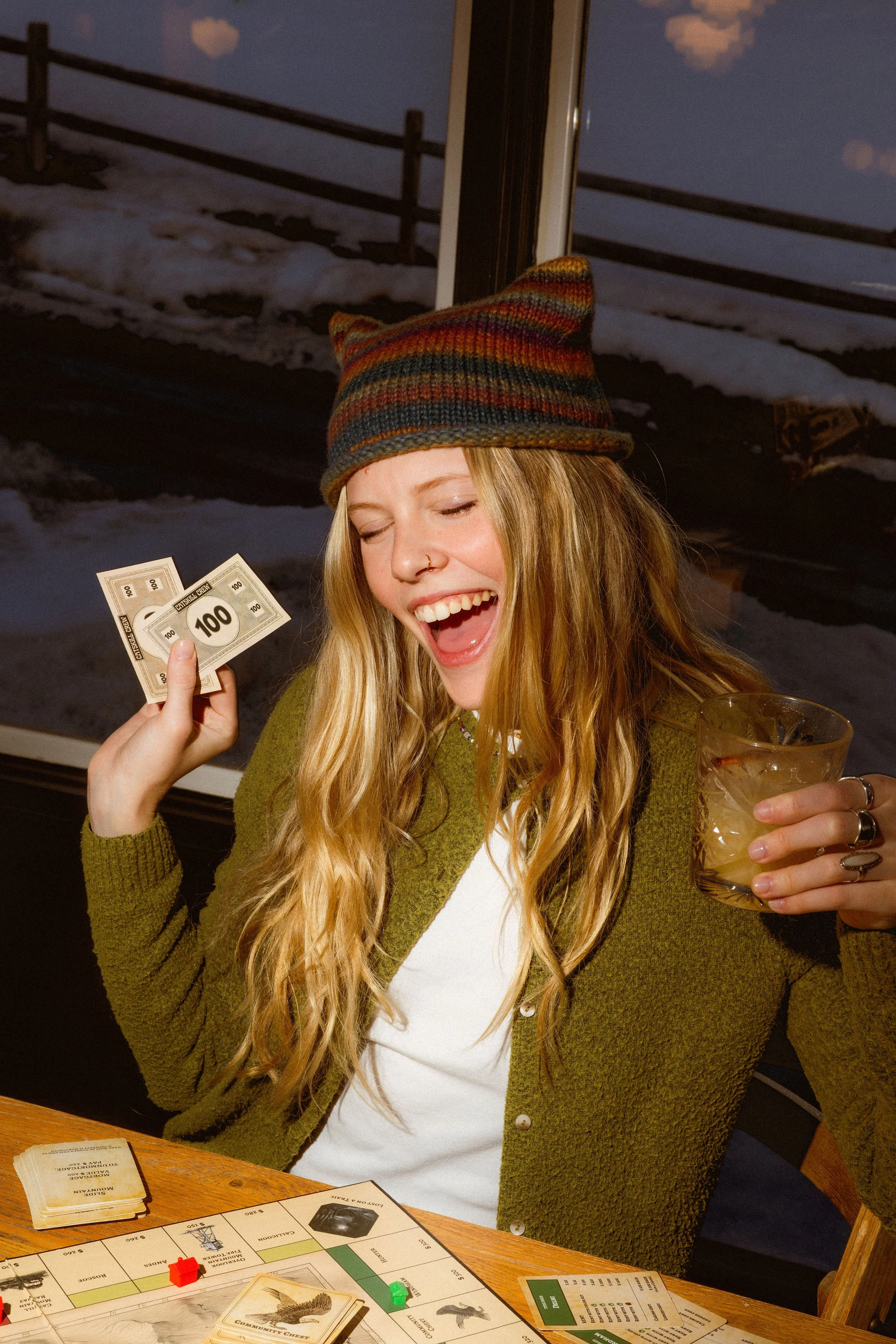 A smiling woman with long blonde hair and a nose ring wears a multicolored knit cap and a green cardigan. She holds up Monopoly money and a glass of a yellowish drink while playing a board game at a wooden table. There is a game board with cards, tok