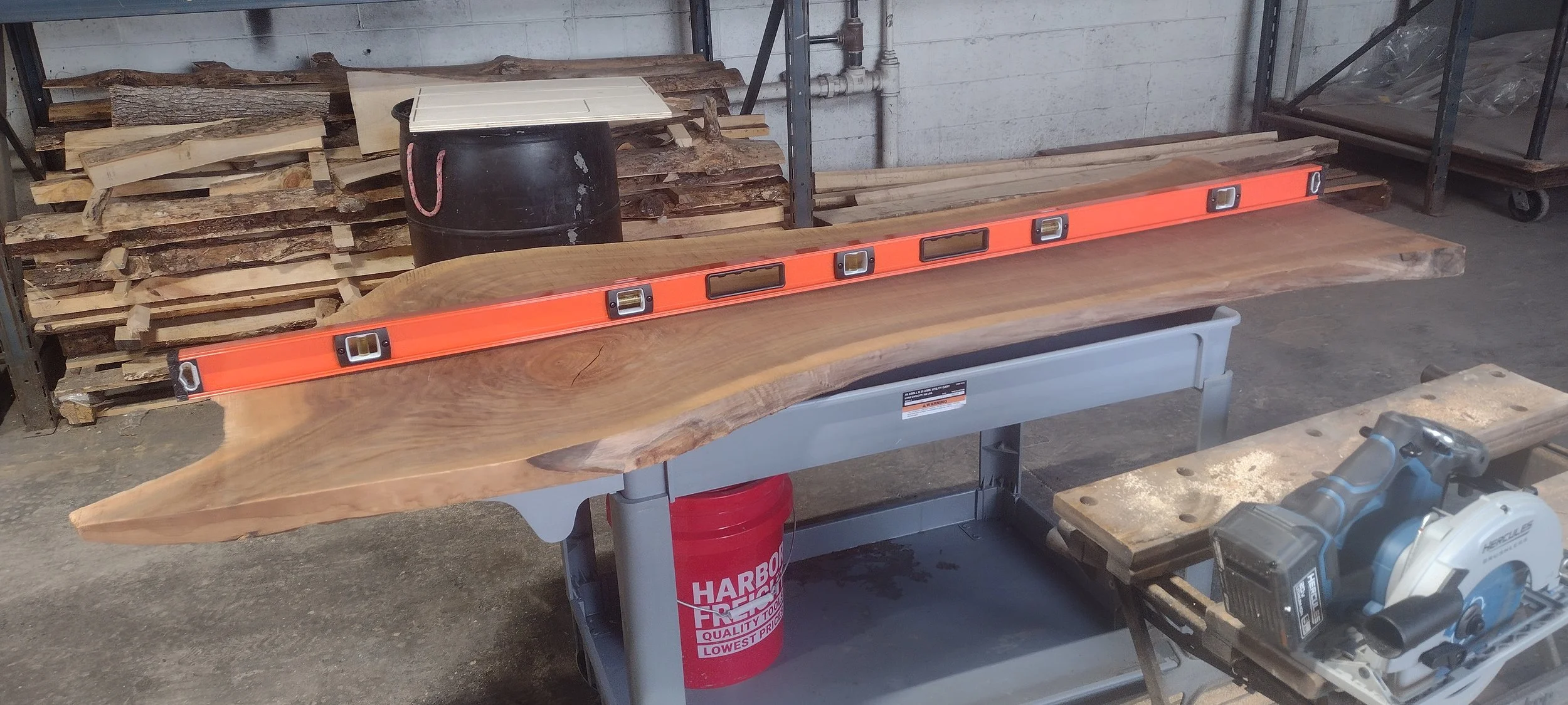 Live edge wooden slab on a workbench with an orange level on top, surrounded by woodworking tools and lumber in the background.