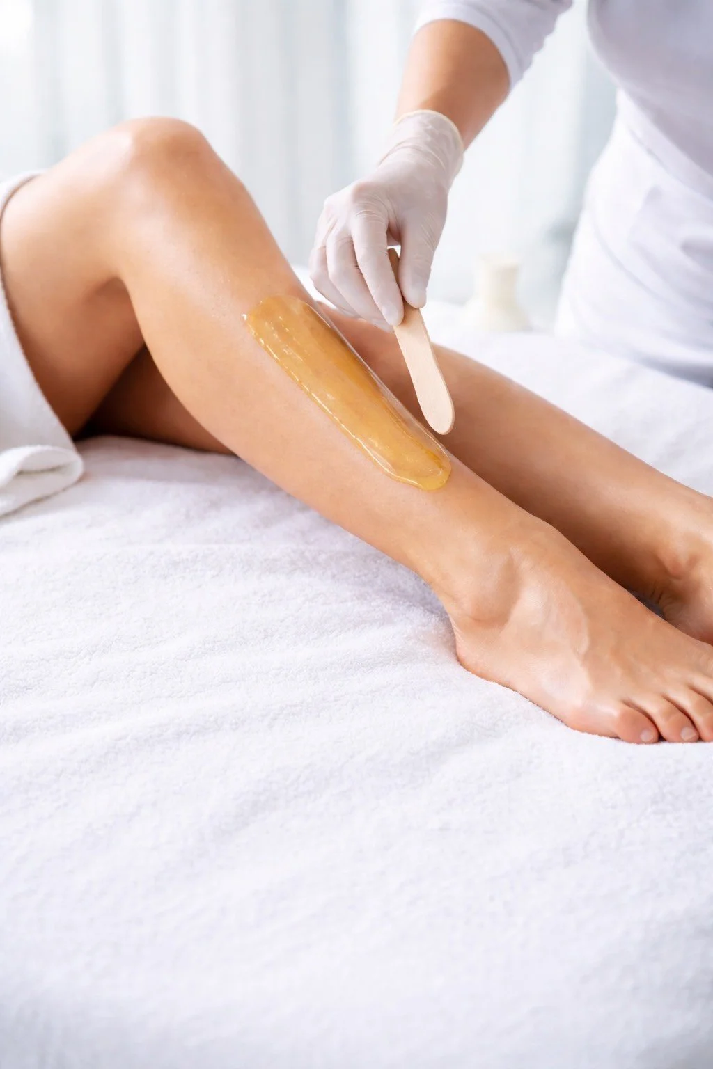 A person receiving a waxing treatment on their leg, with wax applied and a wooden spatula in hand from the esthetician.