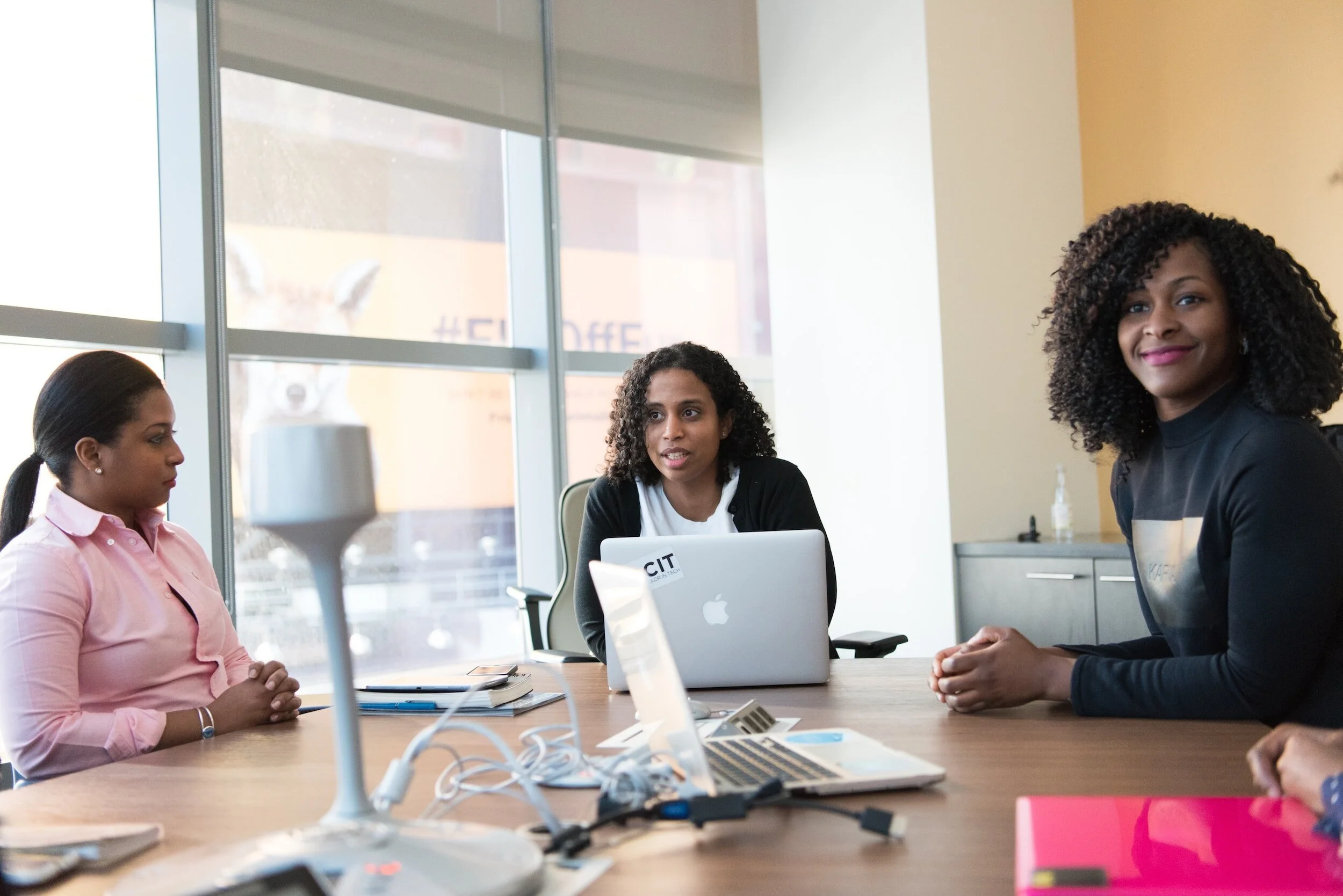 Three women seated around a conference table in a modern office, engaged in a discussion. One woman has curly hair and is speaking while the other two listen and smile. The table has laptops, notebooks, and chargers.