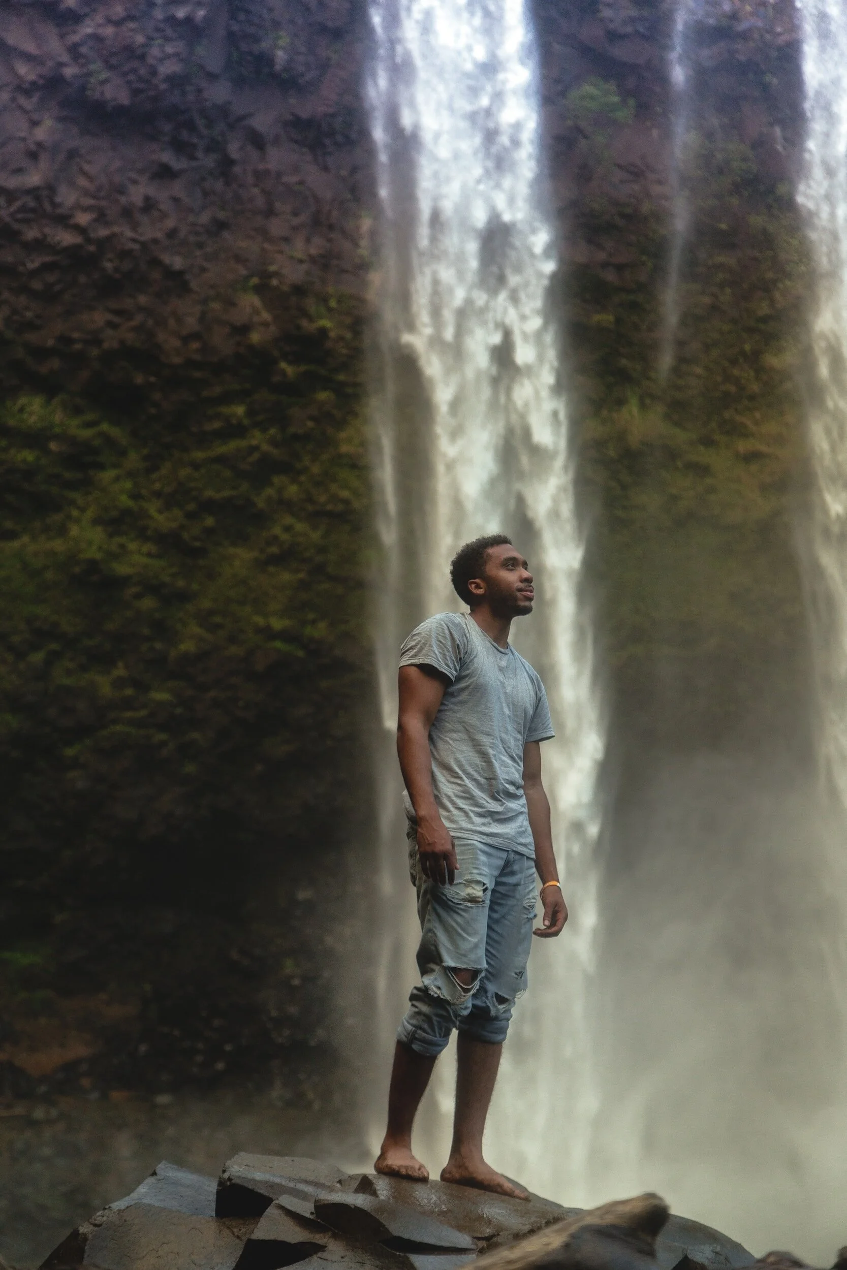 A man standing barefoot on rocks in front of a waterfall with water cascading down a cliff behind him.