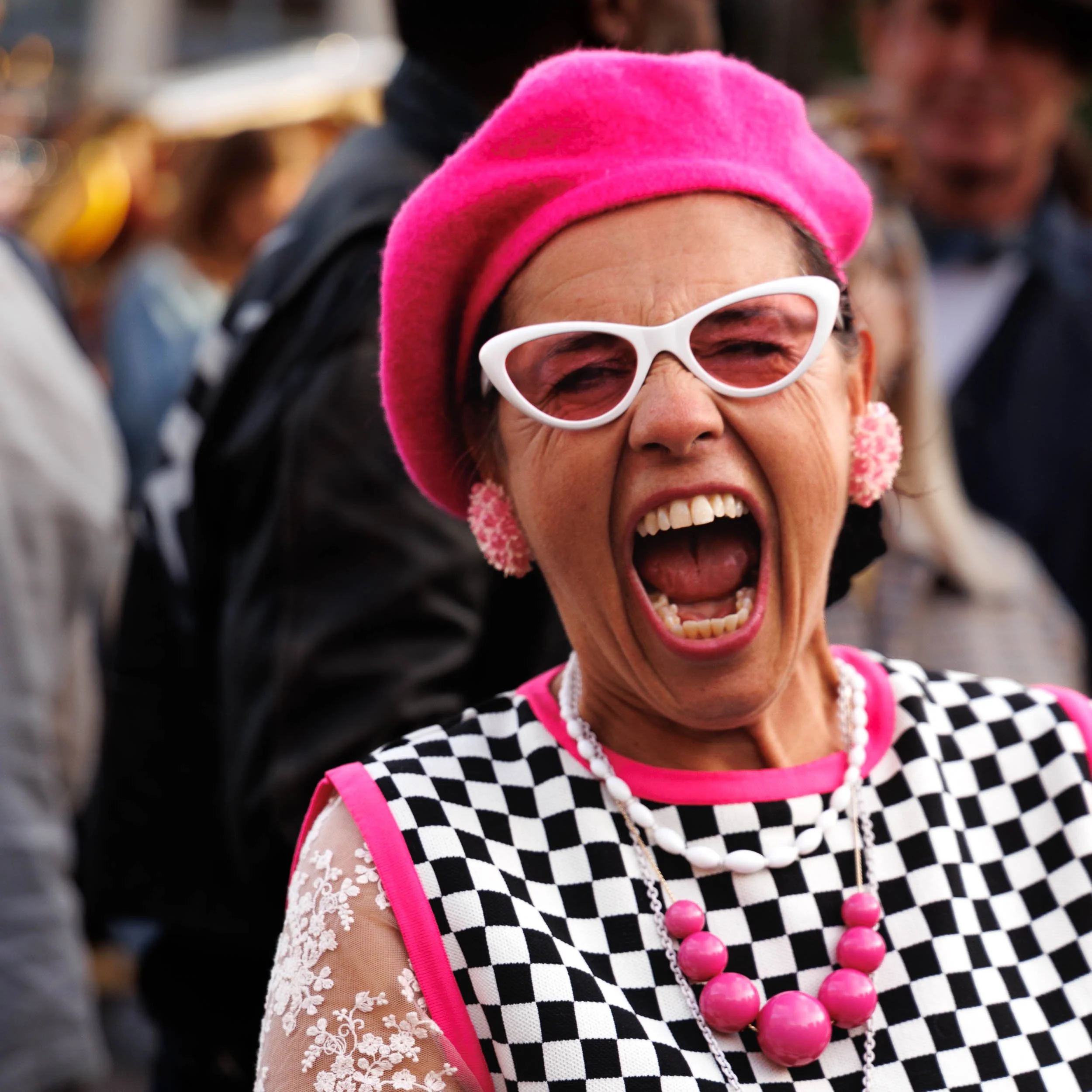 Street portrait in colour of an attractive woman laughing hilariously at the camera. She is wearing a vintage/mod/ska style black and white check dress a pink beret and coordinating sunglasses