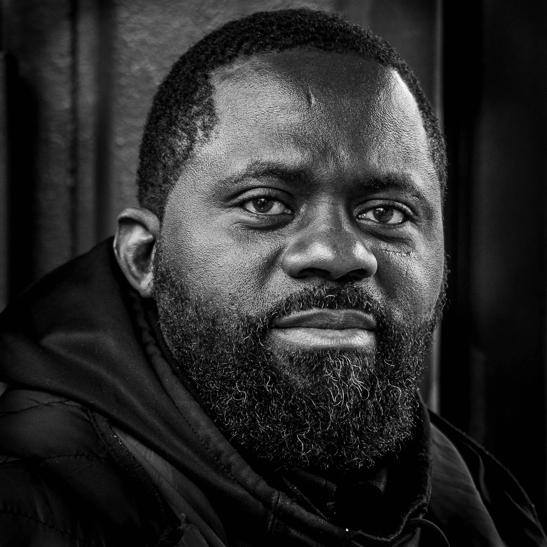 Urban portrait, a tightly cropped headshot of an engaging looking bearded black man, who is looking straight into the camera. image is black and white high contrast
