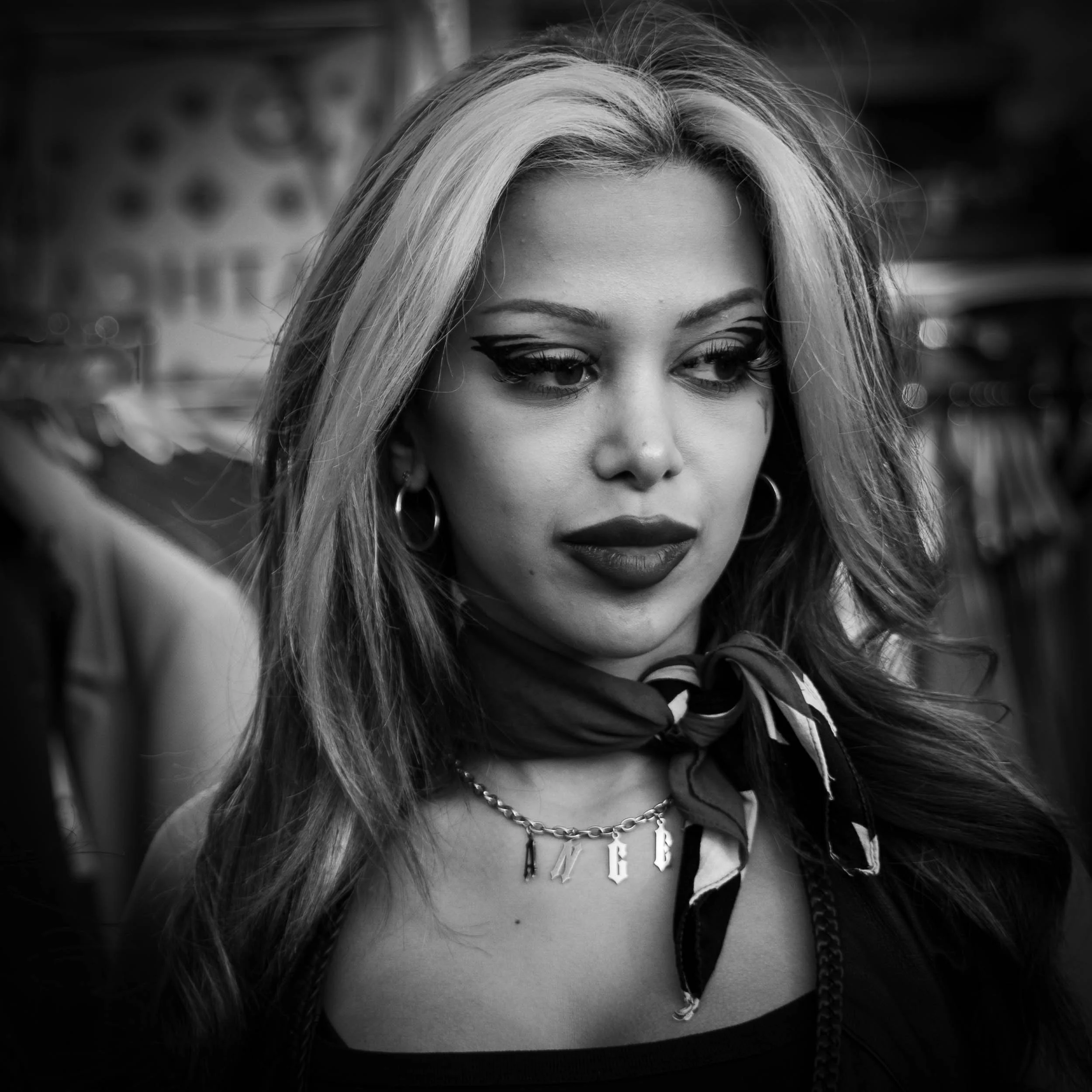 Street portrait of a beautiful young woman, with long hair falling onto her shoulders. She is wearing a leather jacket and has a neckerchief. She is gazing wistfully to the right. A vignette to accentuate the subject in a black and white high contras
