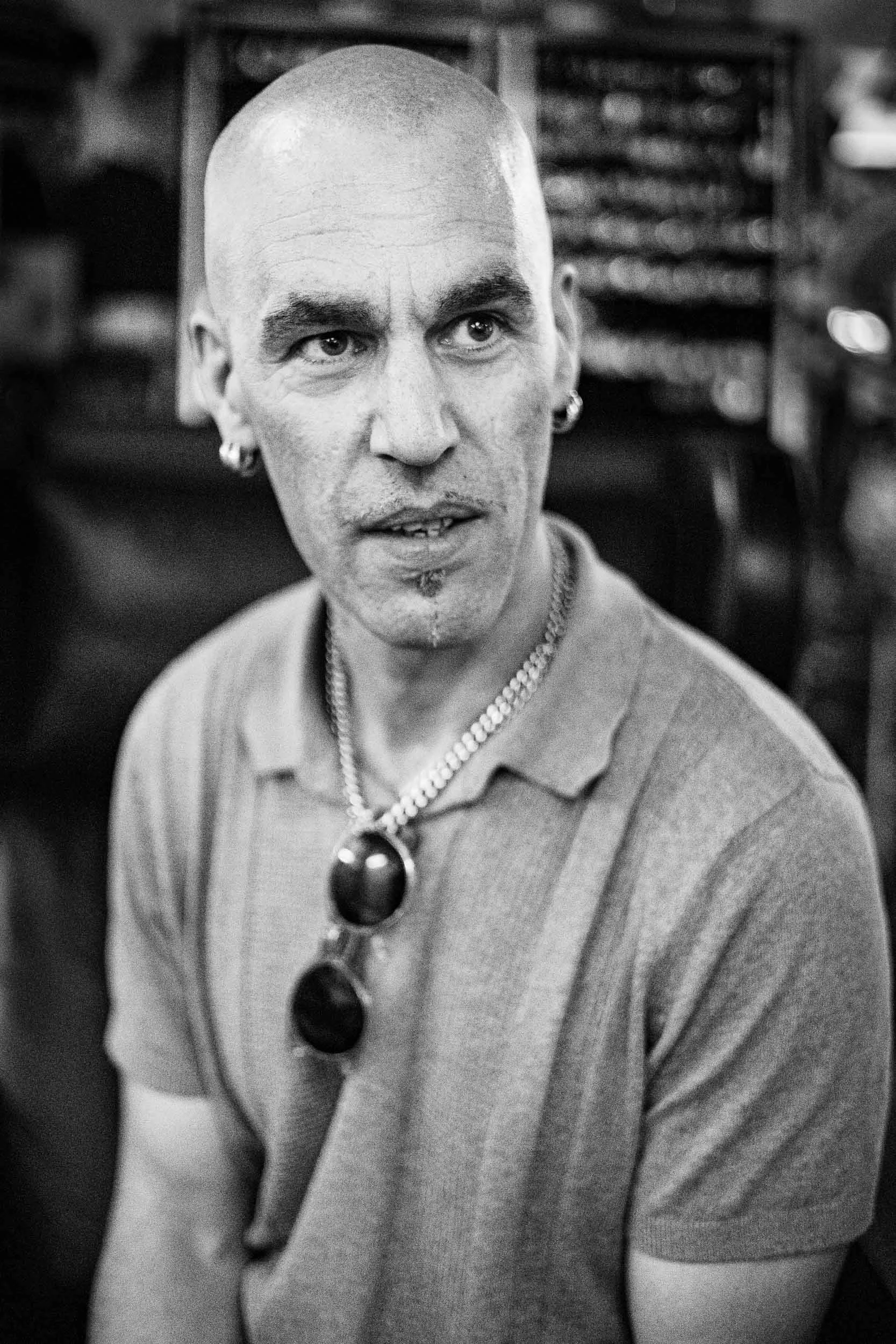 Street portrait of a seated bald man looking to the right, wearing a polo shirt with sunglasses tucked into the collar. Black and white, high contrast with a blurred background.