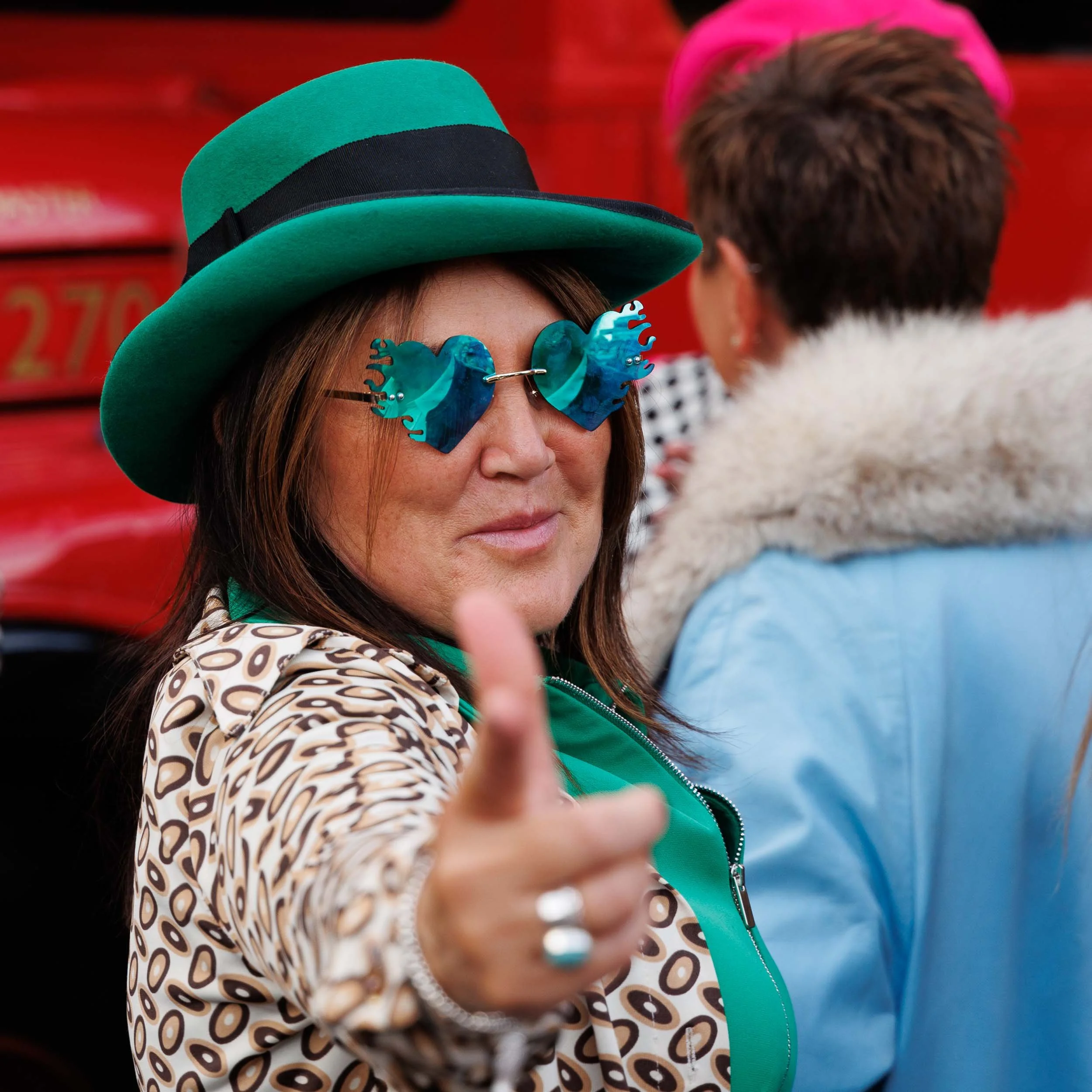 Street portrait in colour of a woman wearing a green fedora with a black ribbon and heart shape green tinted sunglasses and a green shirt looking at the camera while pointing at the photographer.