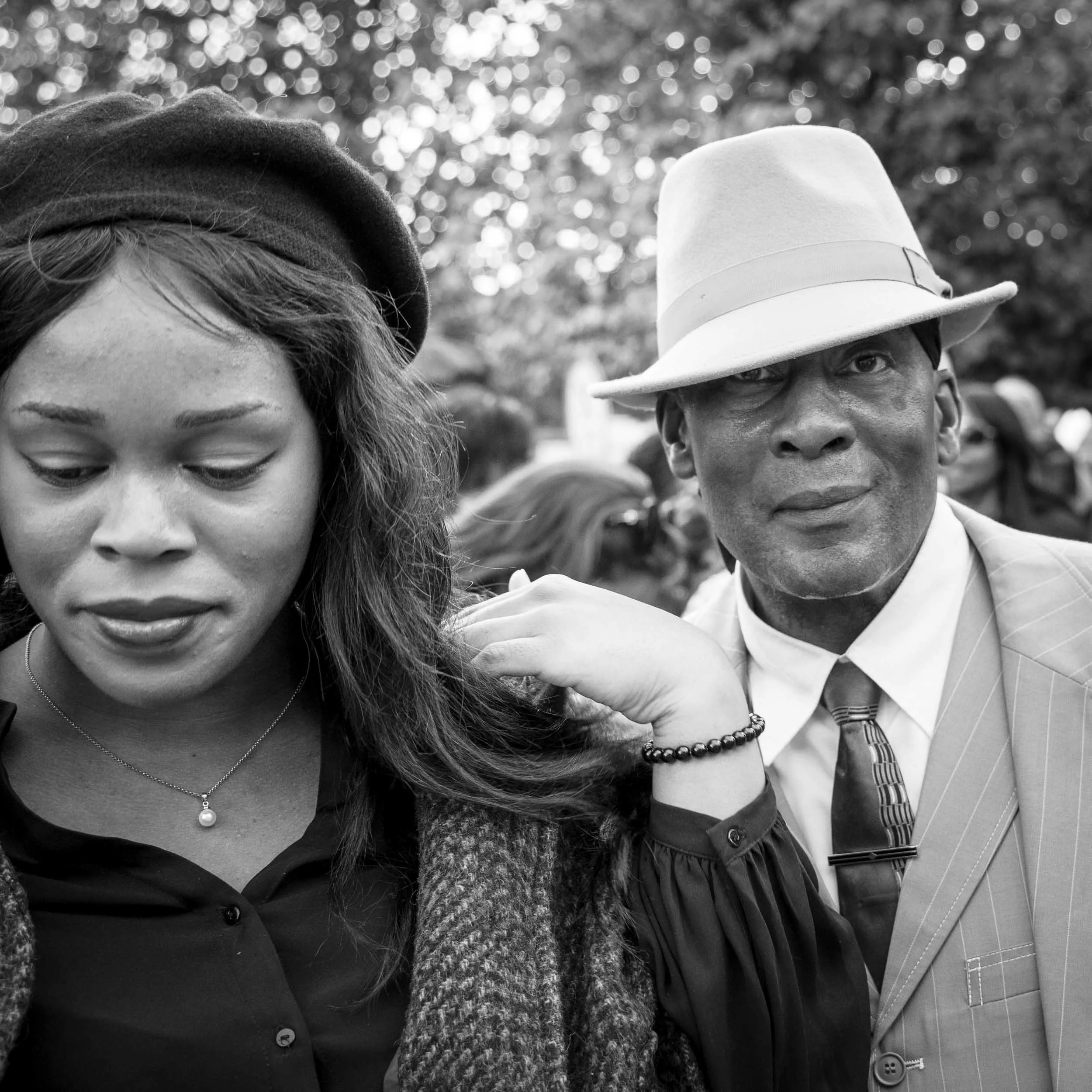 Street portrait of a man and a woman in classic or vintage style clothing. The man is wearing a fedora and is looking into the camera while the woman wearing a beret is looking down shyly. Black and white image