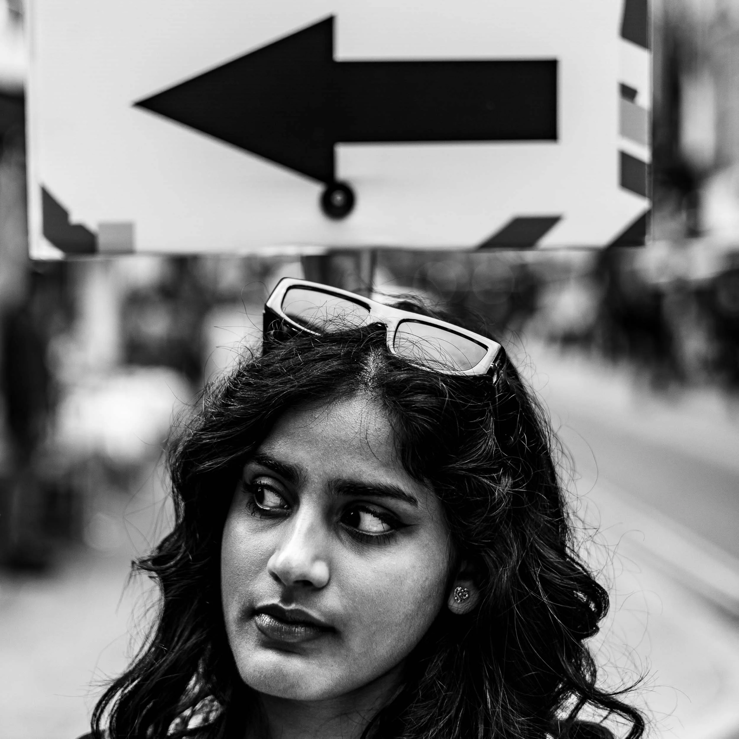 Street portrait of a beautiful young woman with sunglasses perched on her head. close cropped head shot. She is looking to her right underneath a large arrow sign pointing in the same direction as her gaze