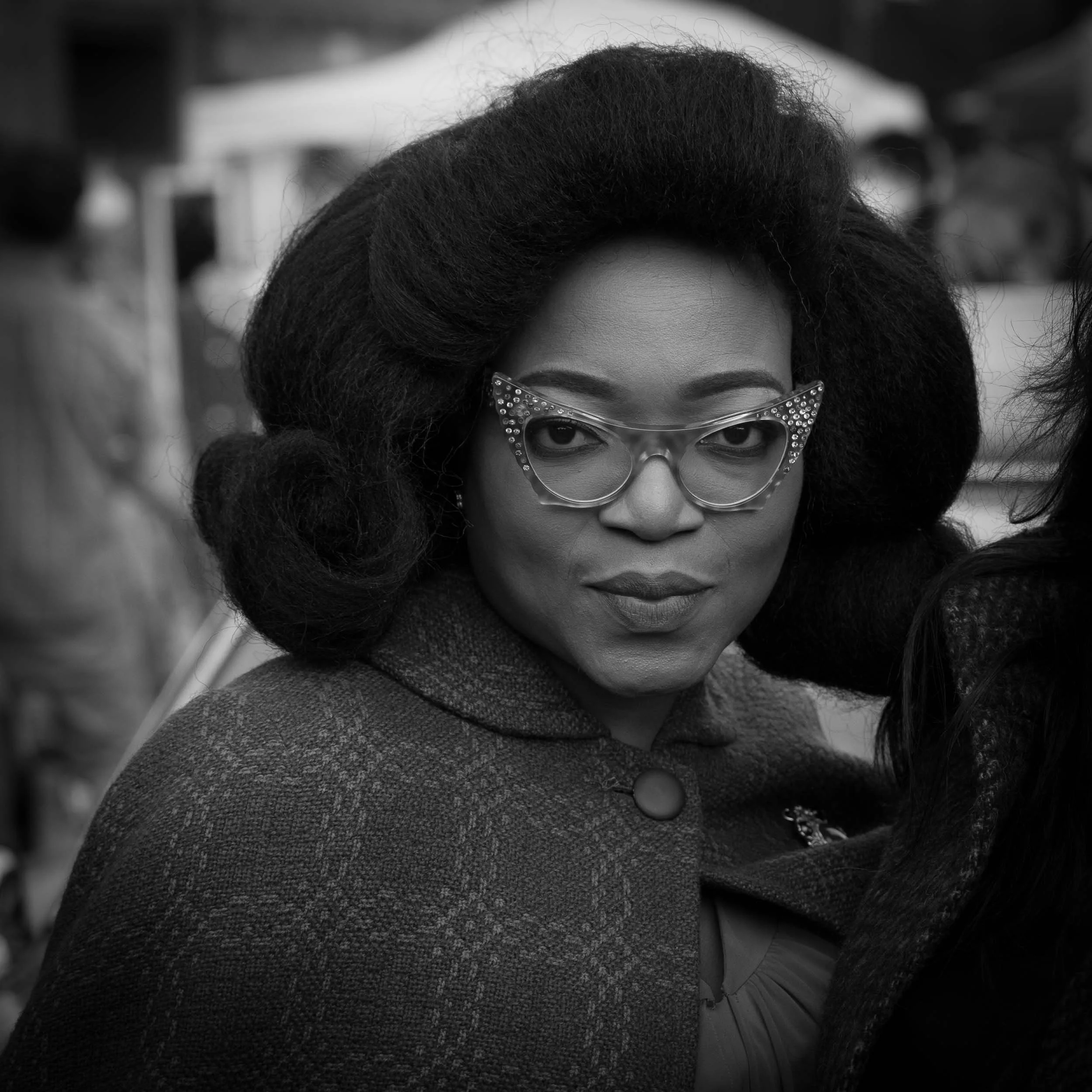 Street portrait of a beautiful black woman, wearing horn rimmed glasses and a bouffant vintage hairstyle, Black and white high contrast image, using a wide aperture for bokeh and blurred background