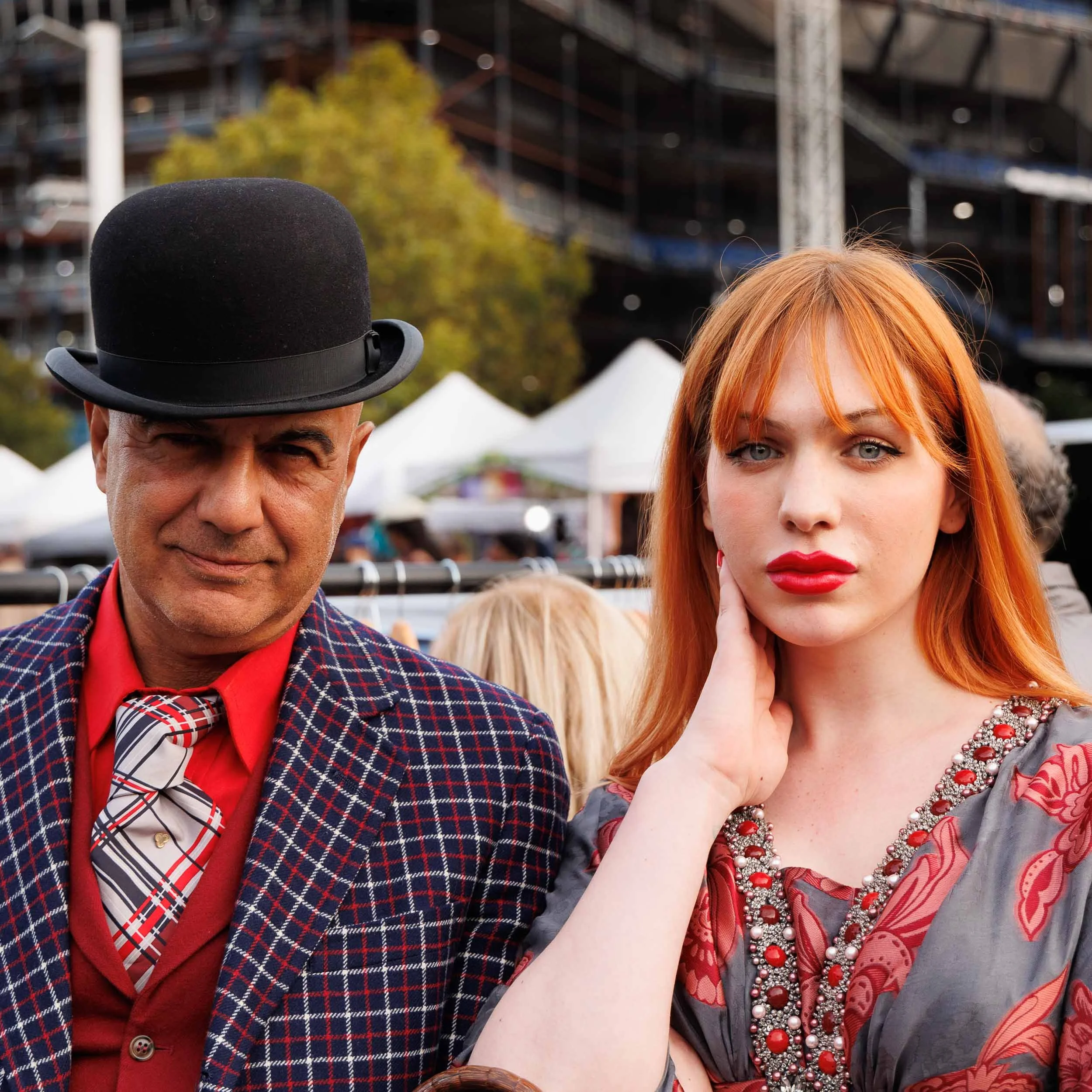  Street portrait in colour of a stylish couple. The man wears a plaid suit, with a red shirt and a bowler hat The red headed woman is wearing a brightly coloured dress and vivid red lipstick her hand positioned to groom her long hair.