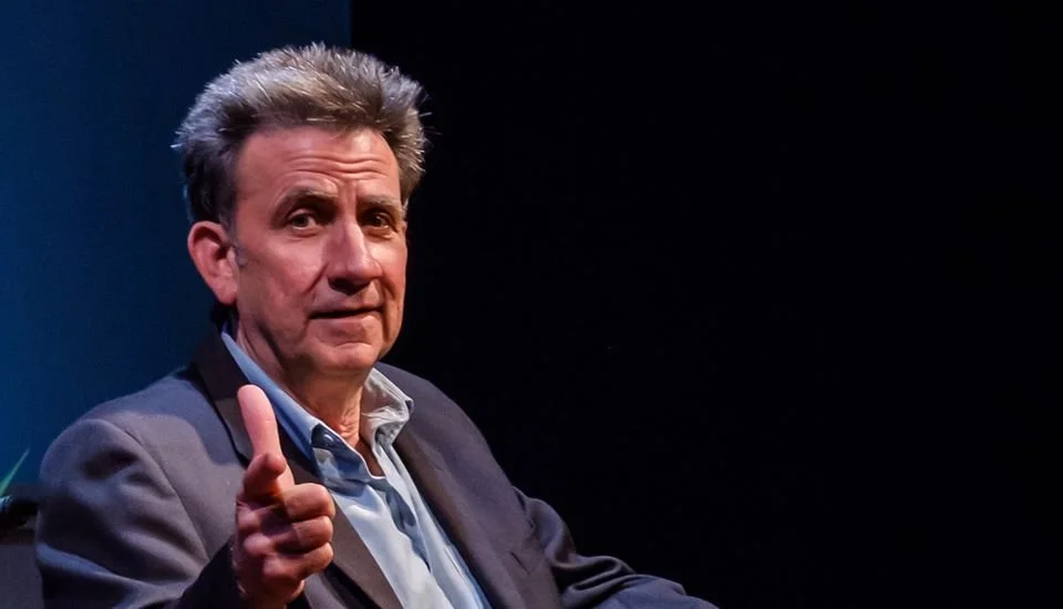 A seated white man in late middle age with salt-and-pepper hair, wearing a suit coat over a dress shirt with no tie, points at the camera, smiling