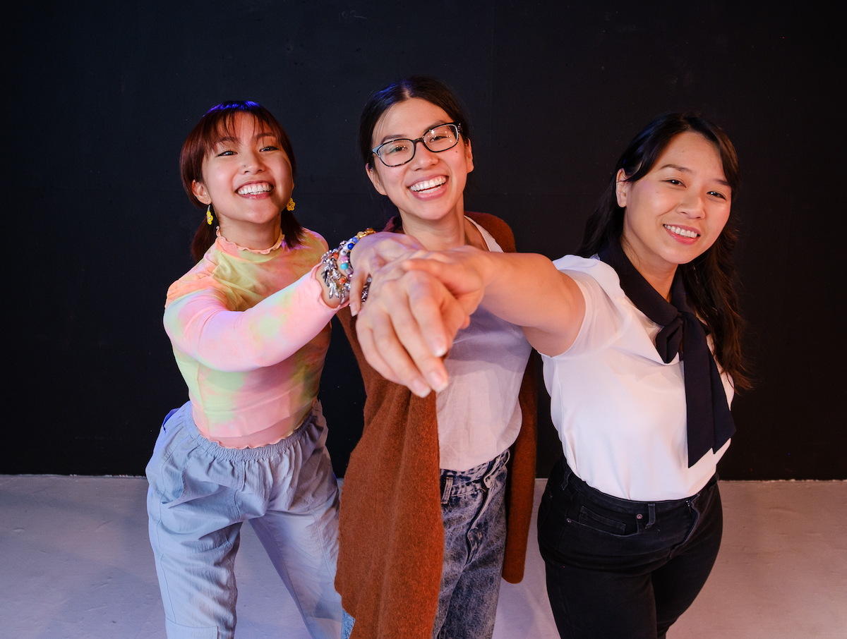 three young Asian women with big smiles, each with one arm stretched out, their hands piled together