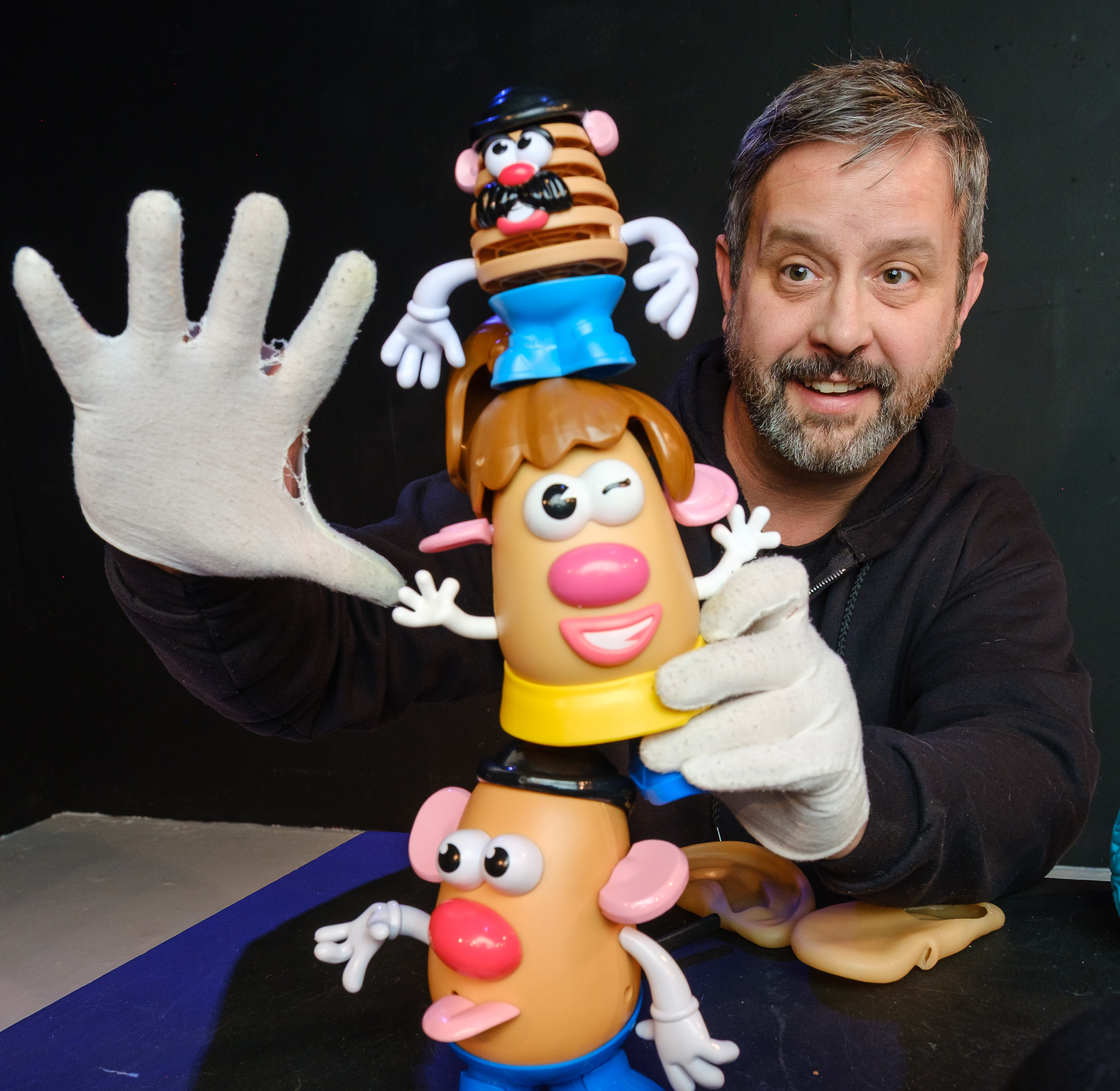 A white man with salt-and-pepper hair, mustache, and short beard, wearing enormous white gloves, is stacking three Mr. Potato Head, dolls on top of each other. He is smiling, and one gloved hand is held palm out to the camera, fingers spread wide.