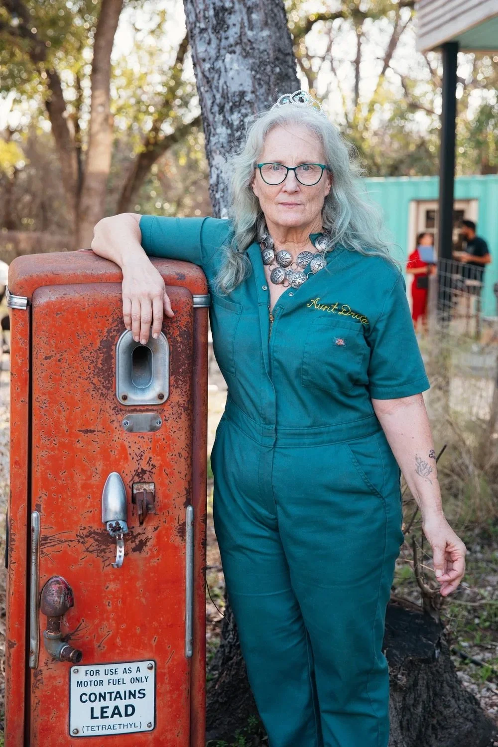 an unsmiling white woman with long white hair in a blue mechanic's coverall leans on a rusting orange gas tank