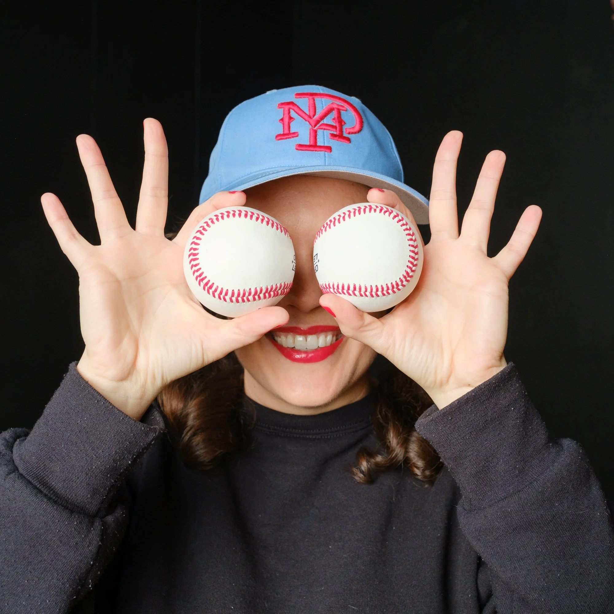 white womsn in blue baseball cap, smiling, holding a baseball in front of each eye, fingers splayed
