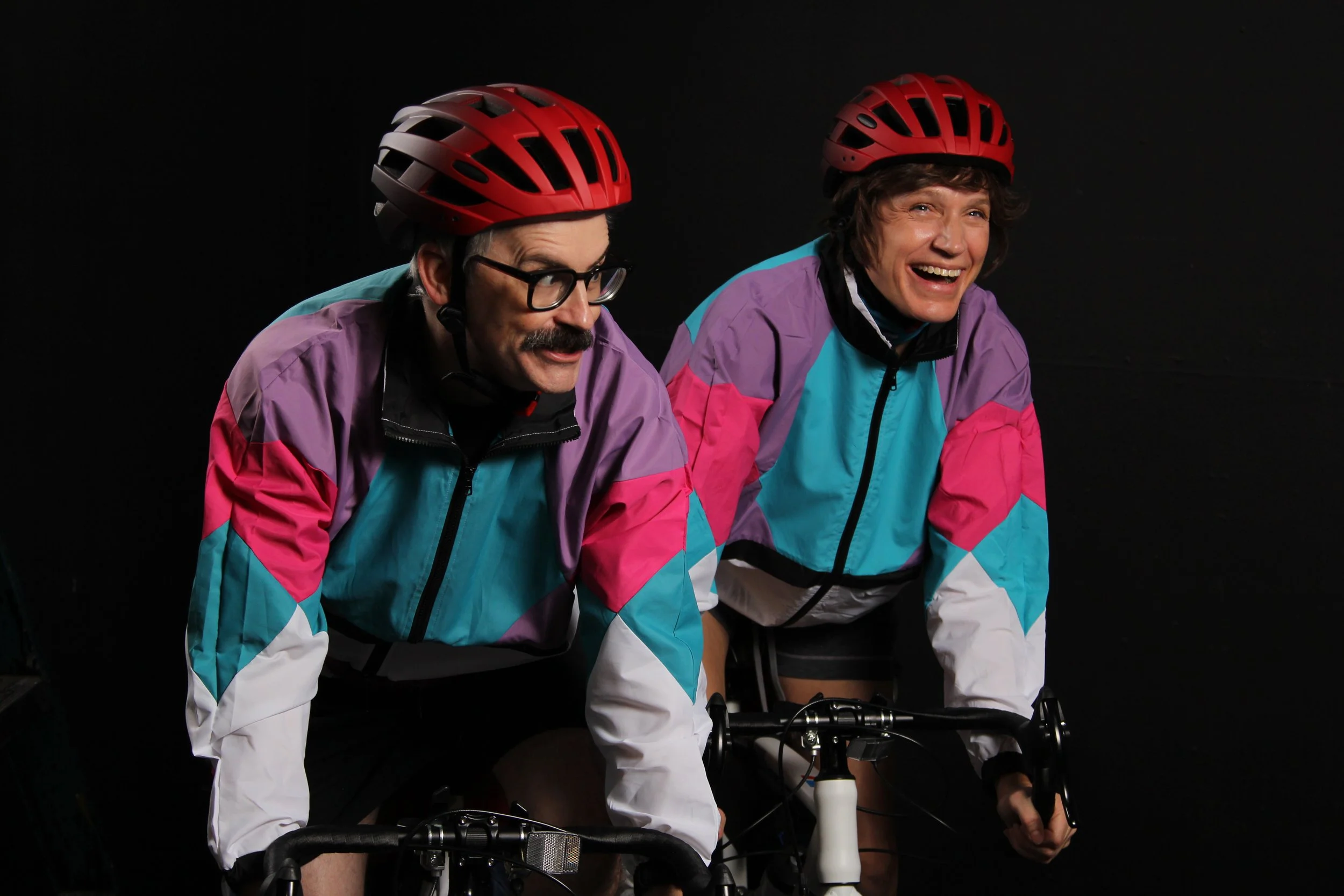A man and a woman on bicycles, wearing colorful windbreakers and bike helpmets. He wears glasses, has a mustache, seems to be saying something funny; she has short hair and is laughing.
