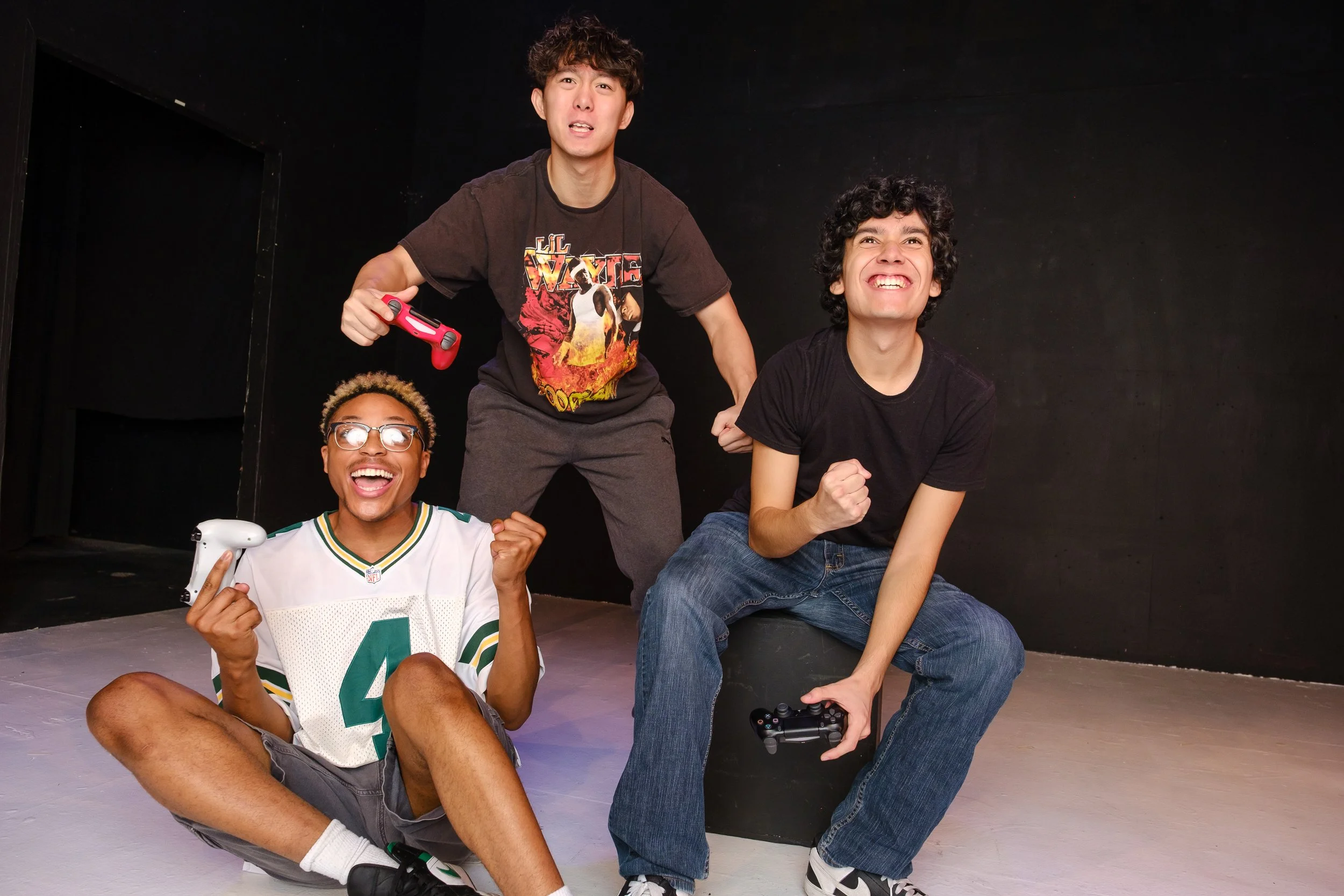 Three young men, perhaps teenagers, holding game controllers: a black man with blond tips and glasses, an Asian man in a Lil Wayne T shirt, and curly-haired Latino man. All three are reacting with delight or intensity to what is happening on screen.