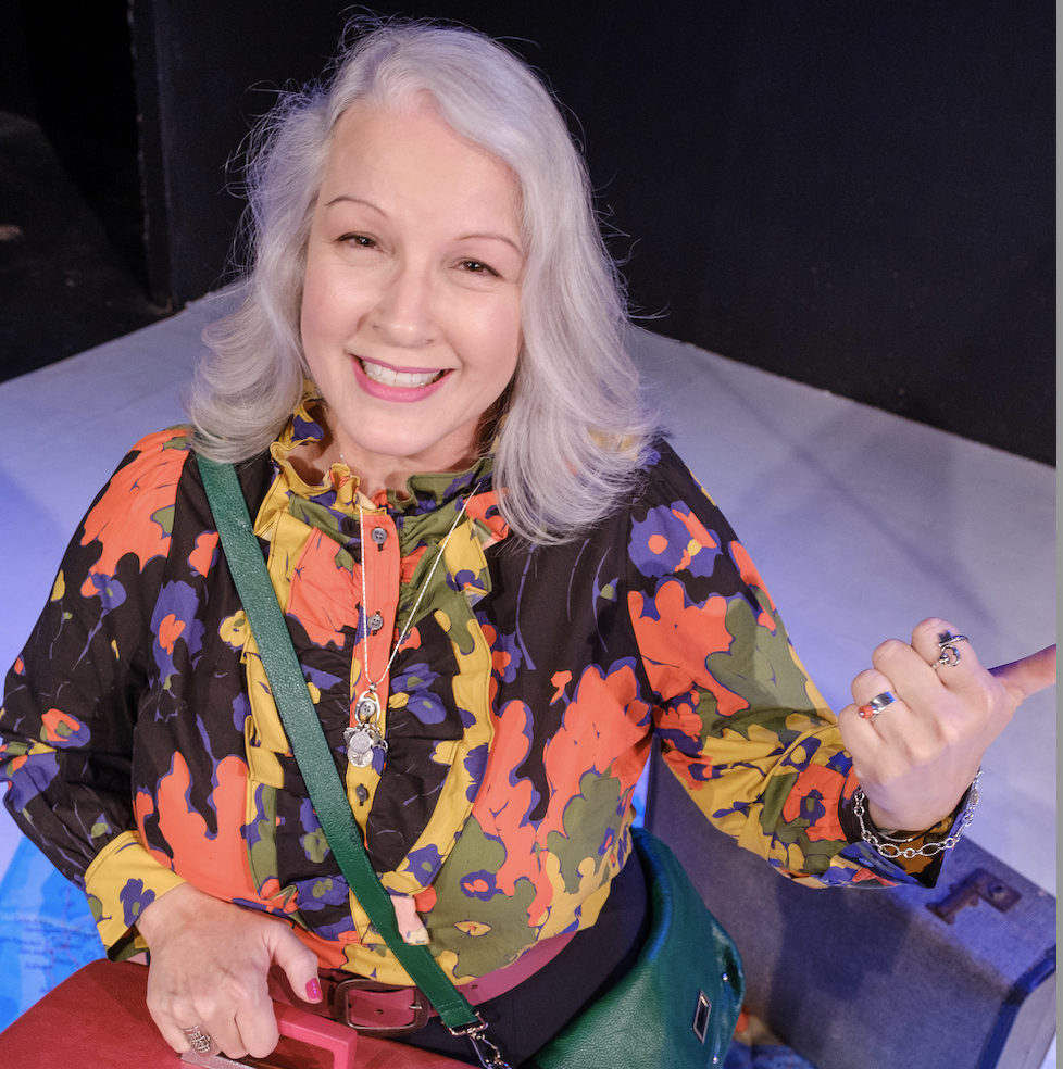 a smiling, pretty white-haired woman in a colorful blouse, her thumb out as if to hitch a ride
