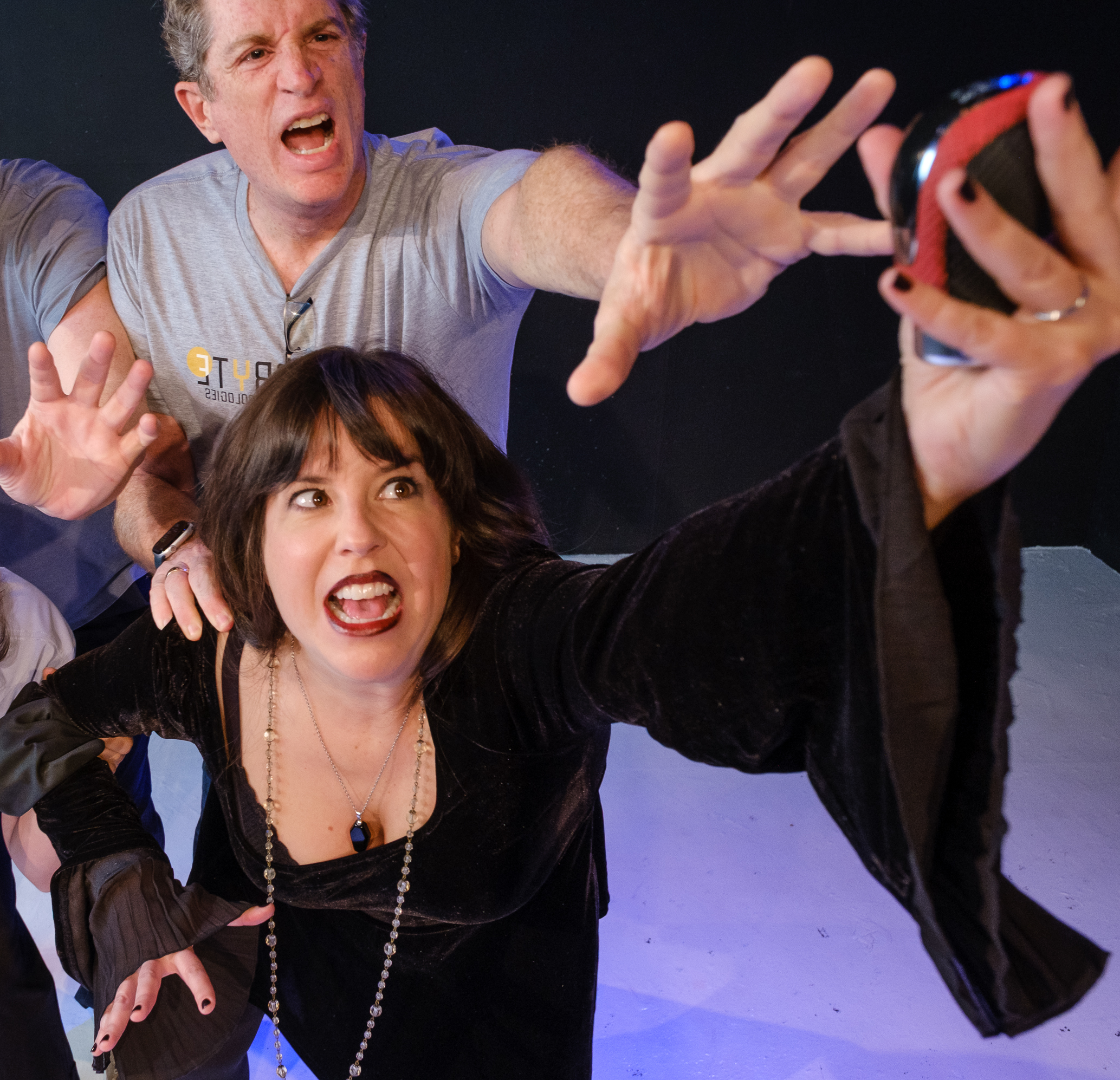 a dark haired white woman in a long black dress with batwing sleeves holds something just out of reach of the two agitated, angry middle-aged white men behind her. Another dark-haired white woman crouches near her, smiling.