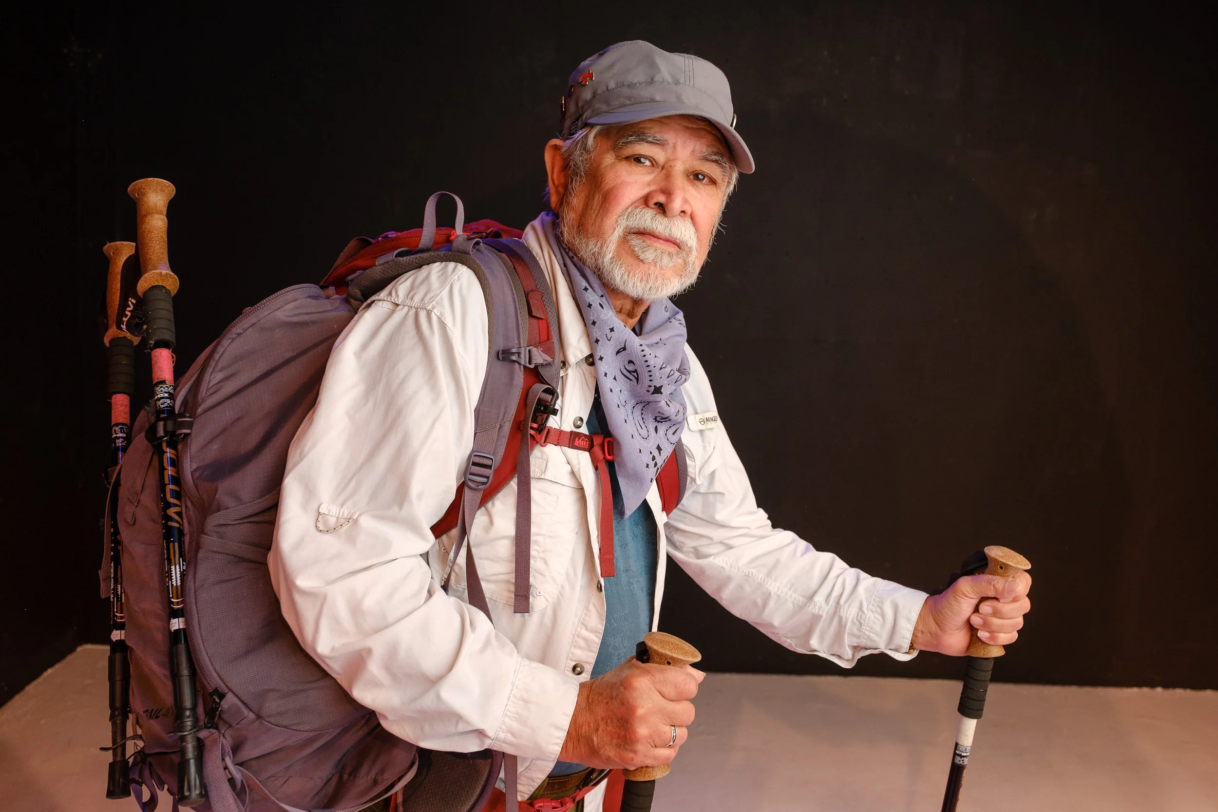 A Latino man with white beard and mustache in a cap and neckerchief wearing a big full pack and walking with two hiking sticks