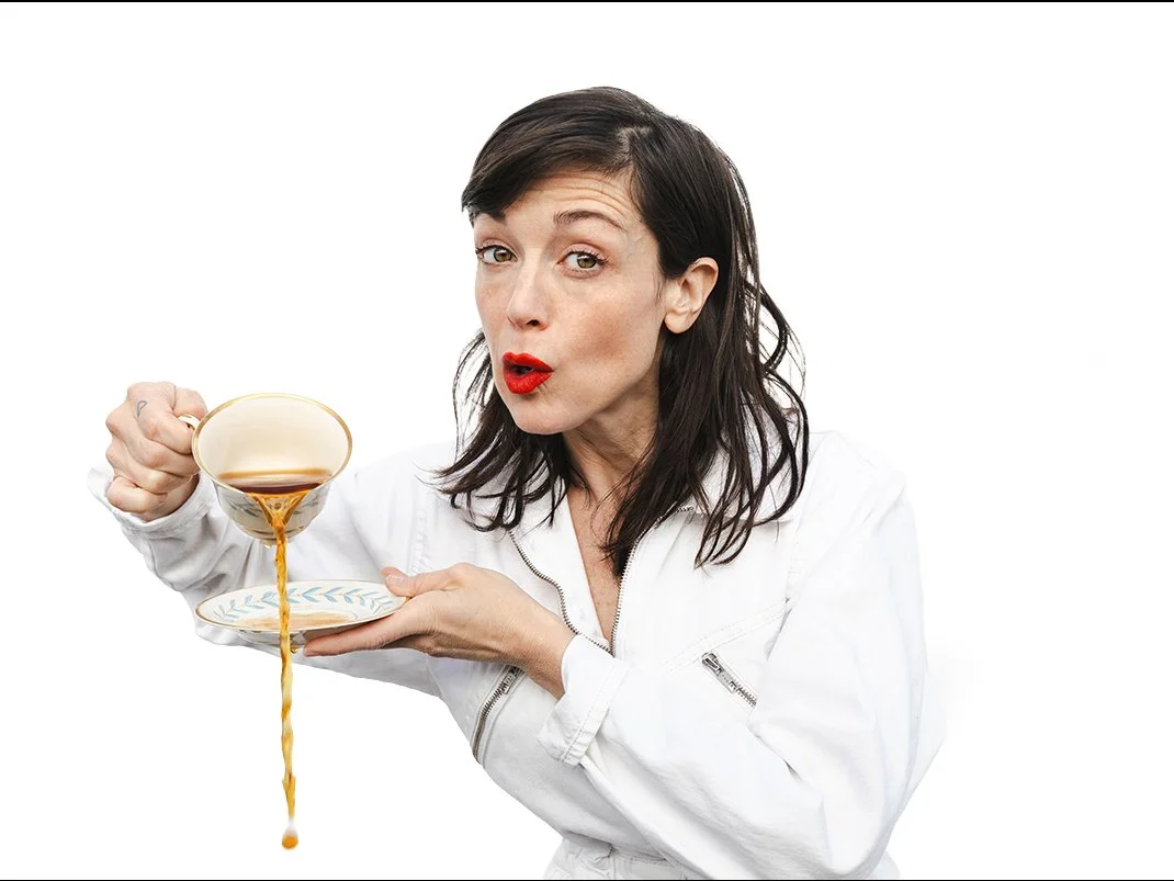 a white woman with tangled dark hair in a white jumpsuit, face in a surprised "Oh!," holding a saucer and pouring tea from a teacup onto the ground