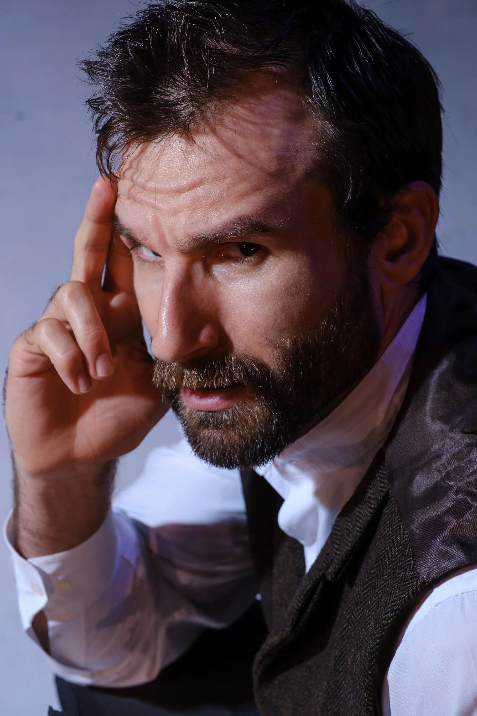 a white man with beard and mustache, hand pressed to temple as if thinking