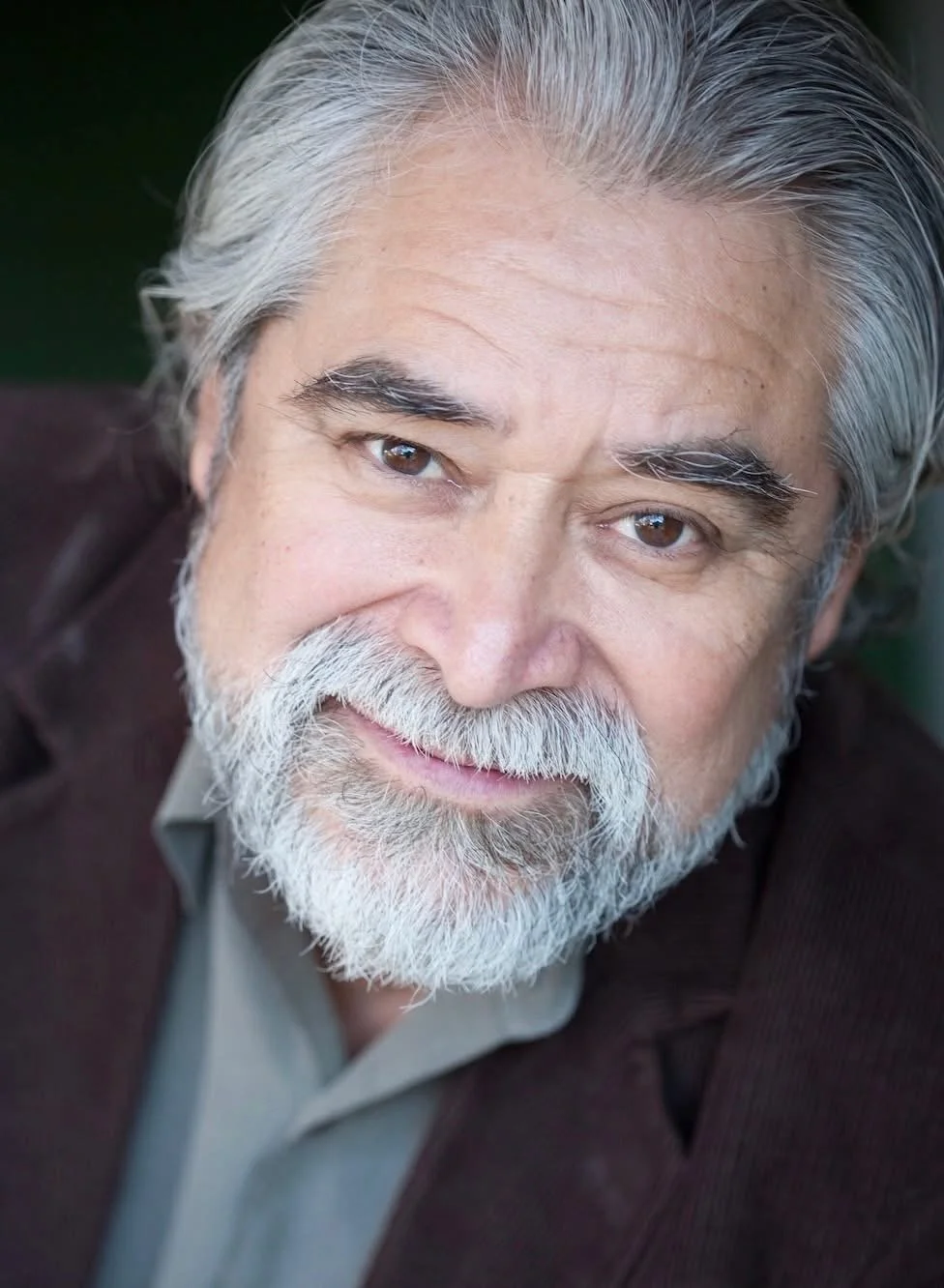 headshot of a faintly smiling older Latino man with silver hair, beard and mustache