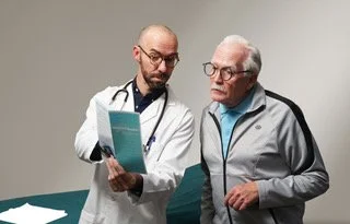 a young balding white male doctor with glasses and facial hair shows a brochure to an elderly white man in athleisure wear