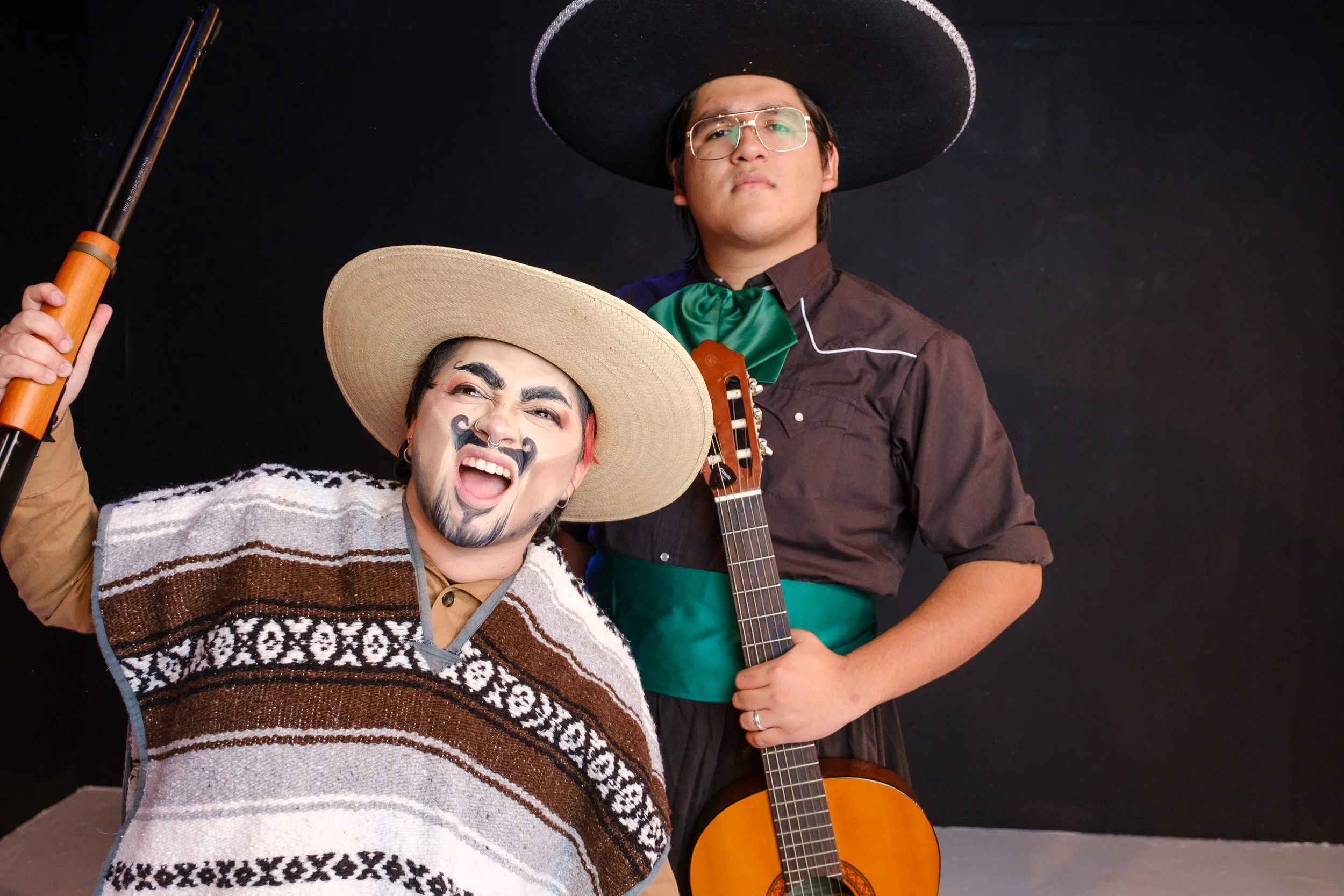 a short, drag-king Pancho Villa raises a rifle, mouth open in a shout. Behind him, a tall Latino in sombrero and glasses, wearing a noble expression, carrying a guitar