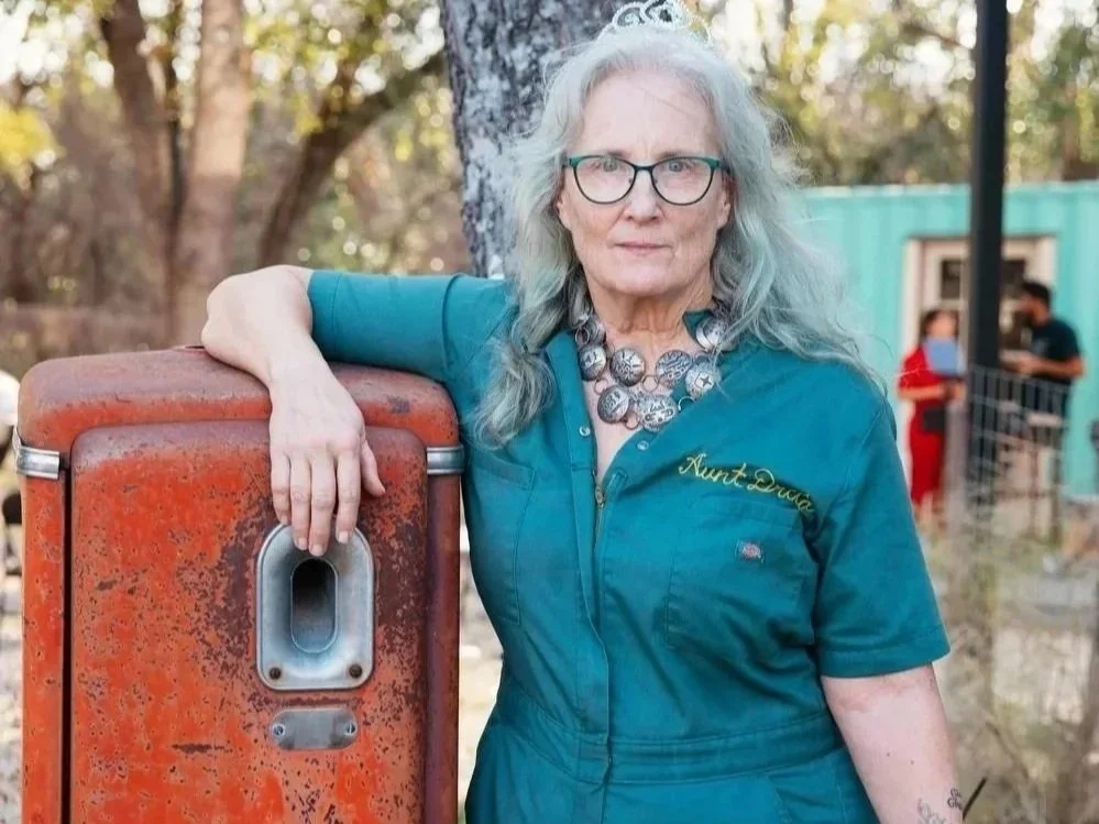 an unsmiling white woman with long white hair in a blue mechanic's coverall leans on a rusting orange gas tank 