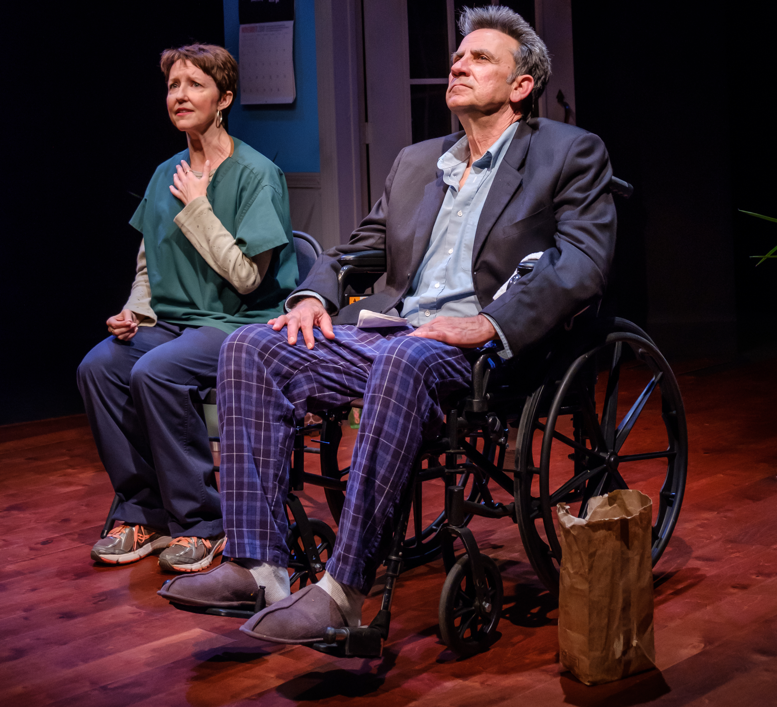 A white man in late middle age with salt-and-pepper hair, wearing a suit coat & a dress shirt with no tie over pajama pants, in a wheelchair; seated beside him a white woman with short dark hair, hand to her throat, moved