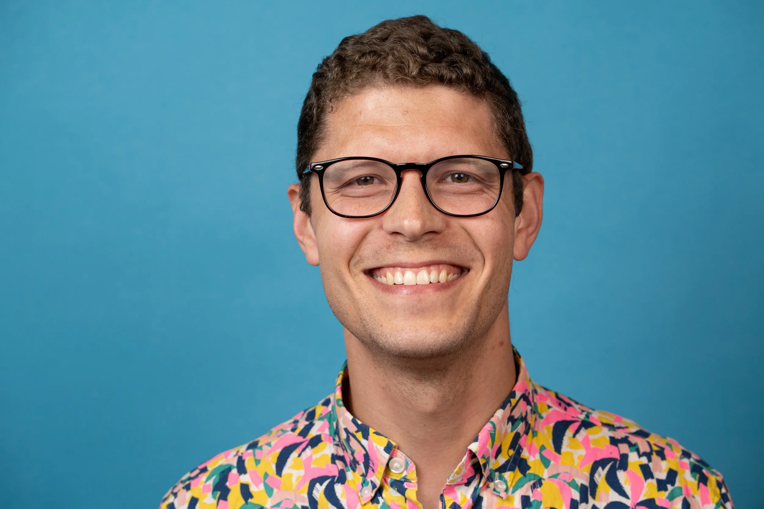 grinning white man with short, curly dark blond hair grins at camera before a bright blue background