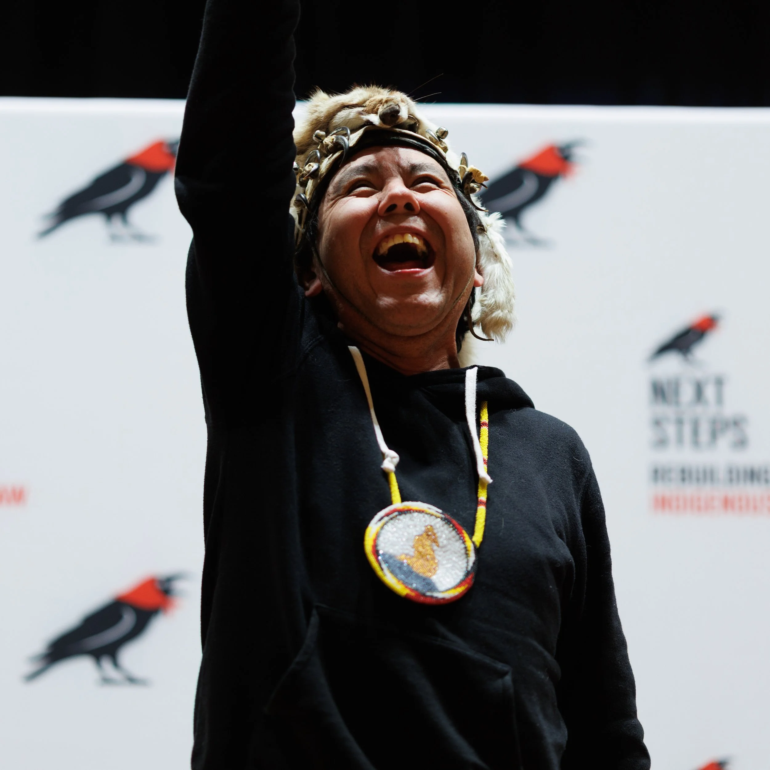 A man wearing a Yak hat and a medal around his neck, smiling and celebrating on stage, with a background displaying the logo of a black bird.